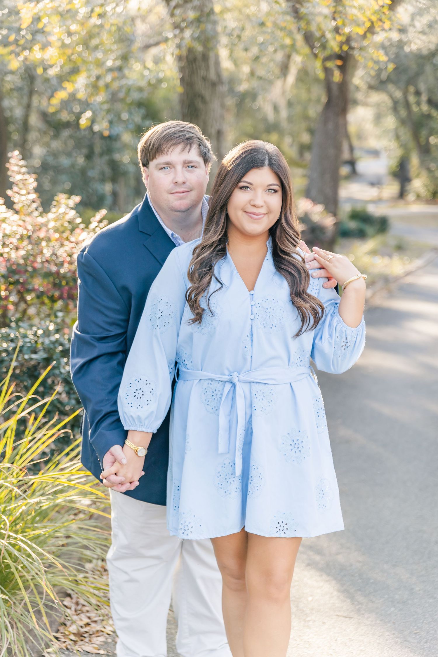 Couple standing together on a wooded path, with her in a light blue eyelet dress and him in navy and khakis.