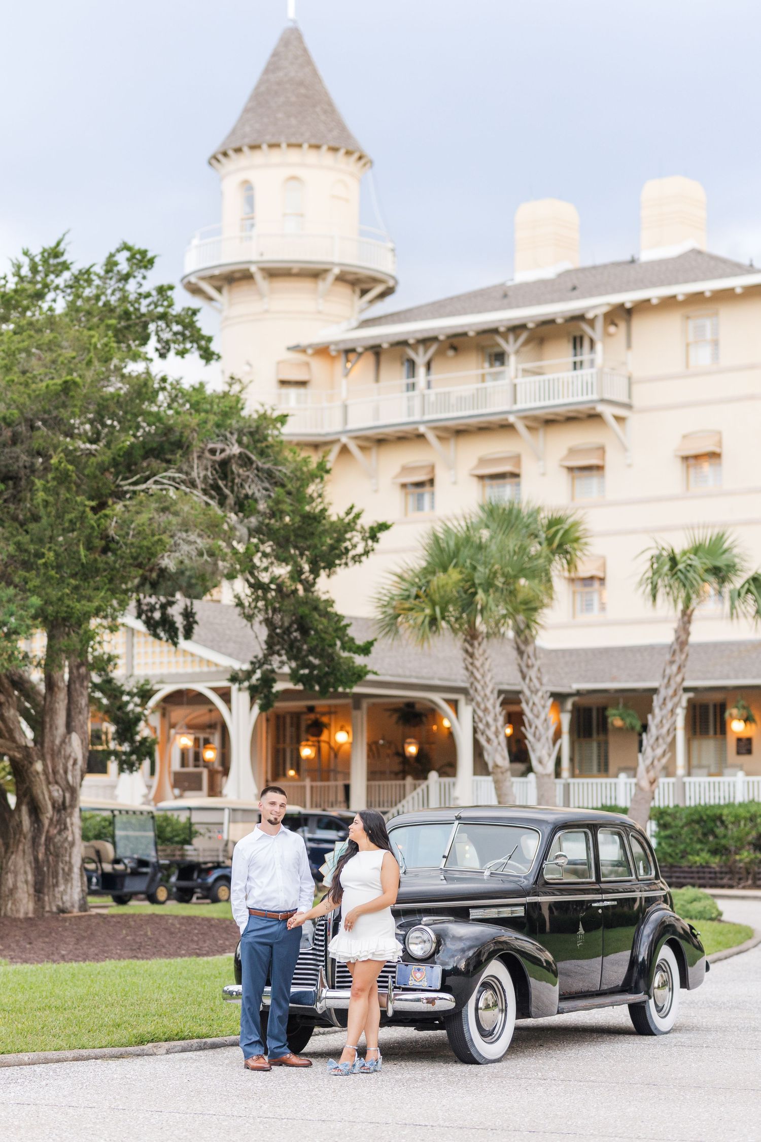 Couple poses beside vintage black classic car in front of historic Victorian yellow hotel with turret.