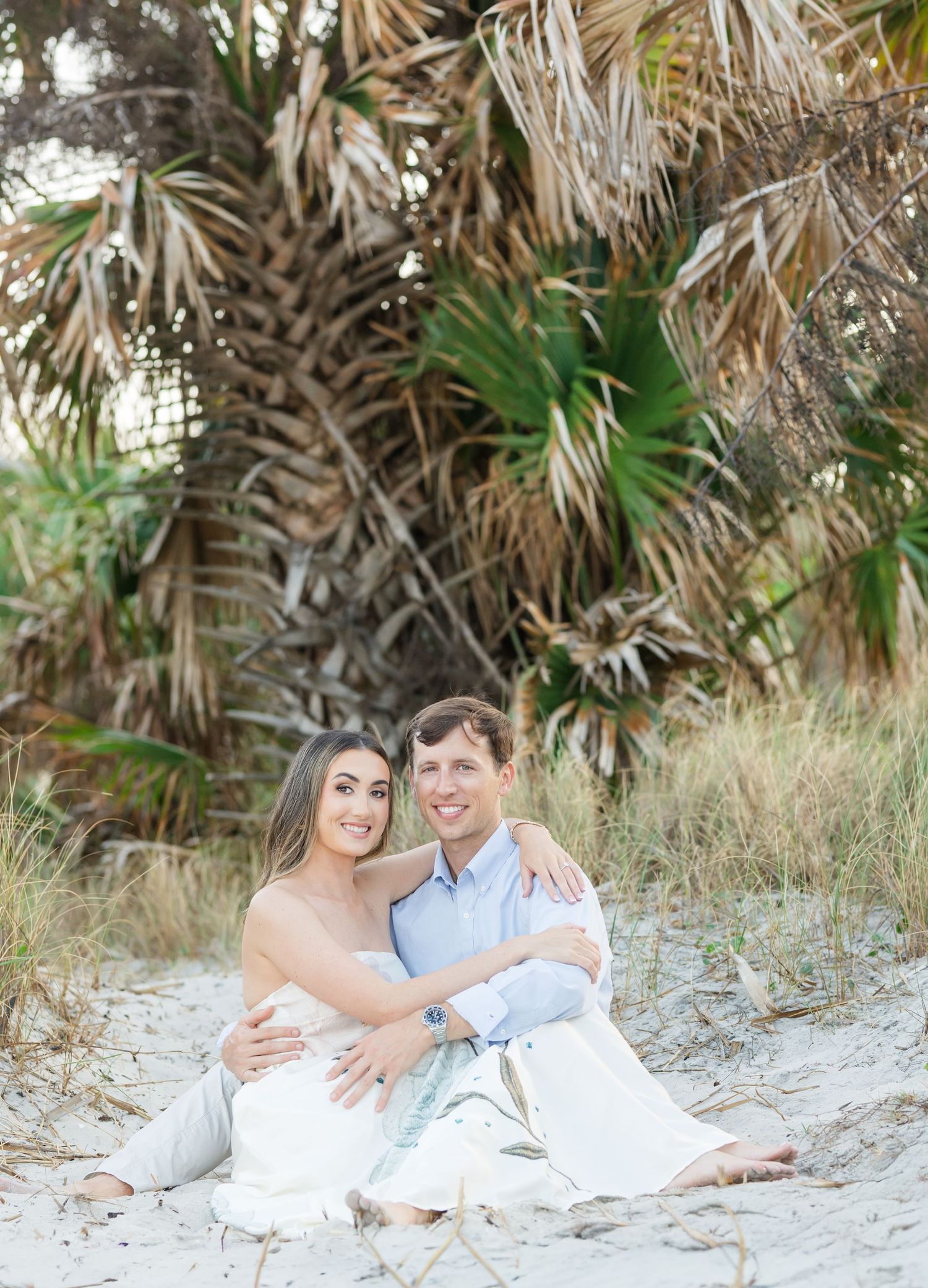 Happy couple sitting together on sandy beach dunes with lush palm trees behind them.