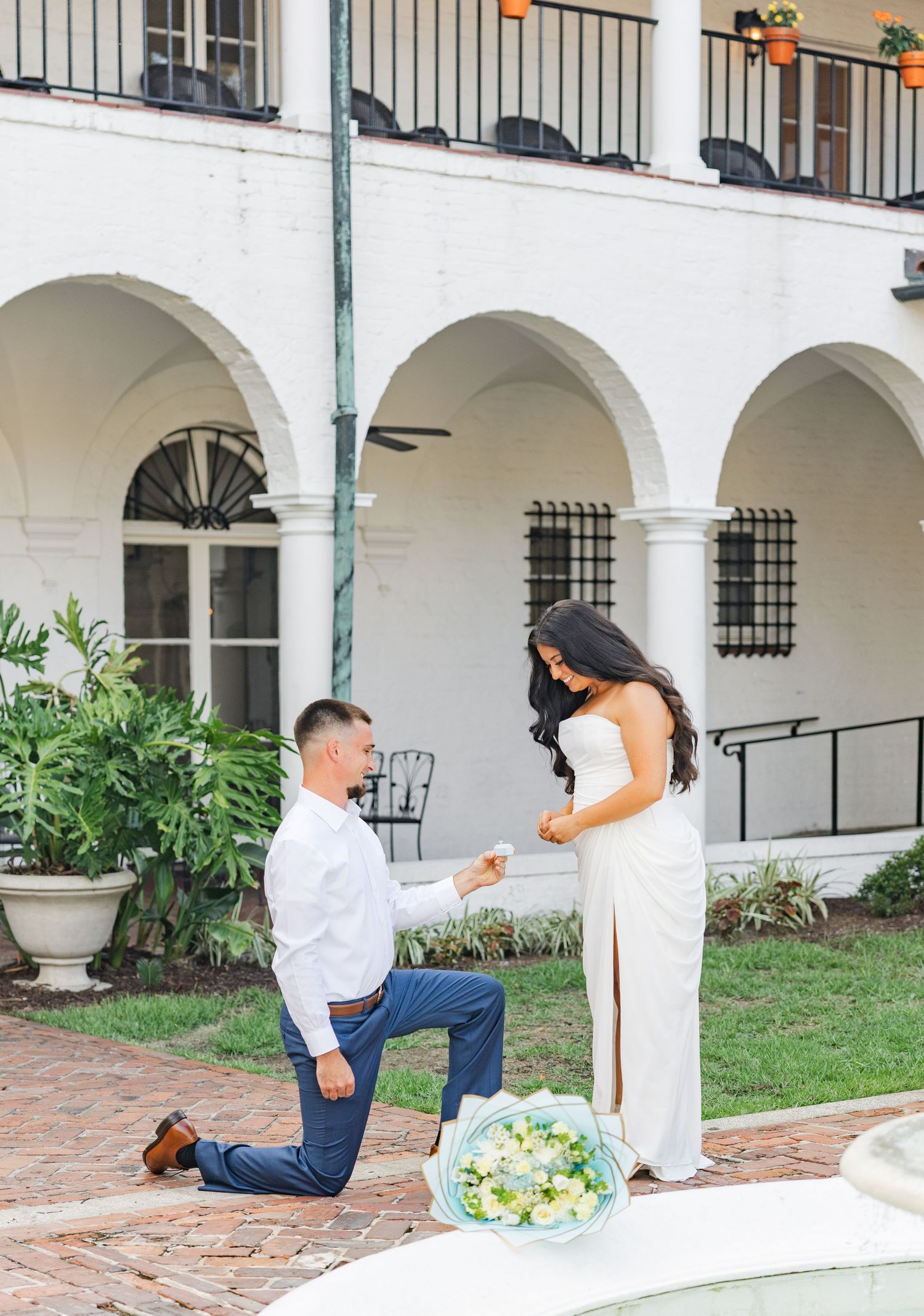 Man proposing on one knee to woman in white dress near fountain with bouquet at elegant white building.