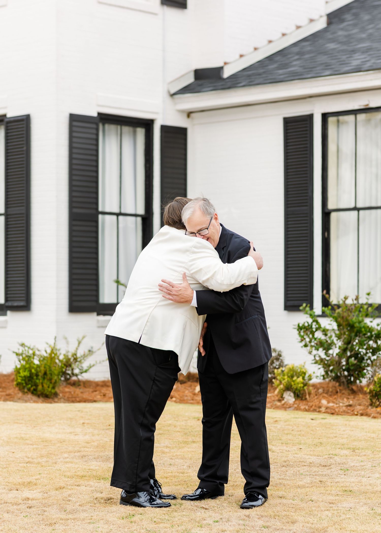 Two men share a heartfelt hug outside a white colonial-style home with black shutters at a wedding.