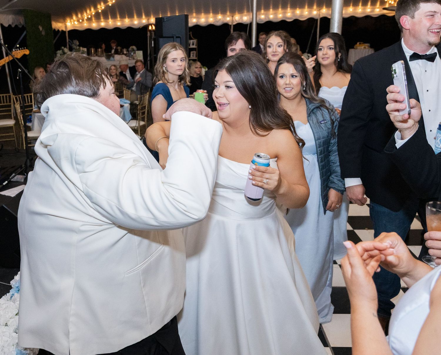 Bride in strapless white gown dancing joyfully with guest in white jacket at lively wedding reception.