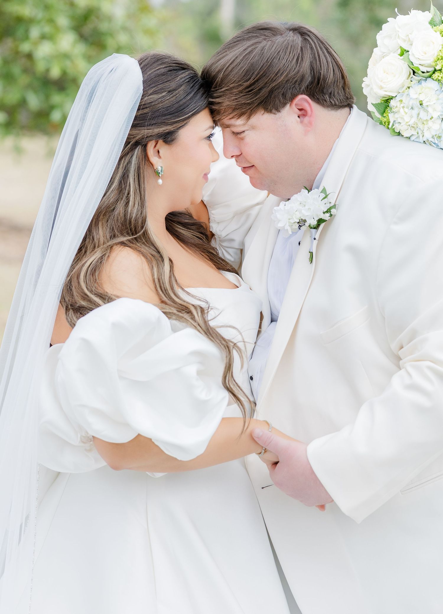 Bride and groom touch foreheads tenderly during outdoor wedding ceremony, she in white gown with veil, he in white tuxedo.