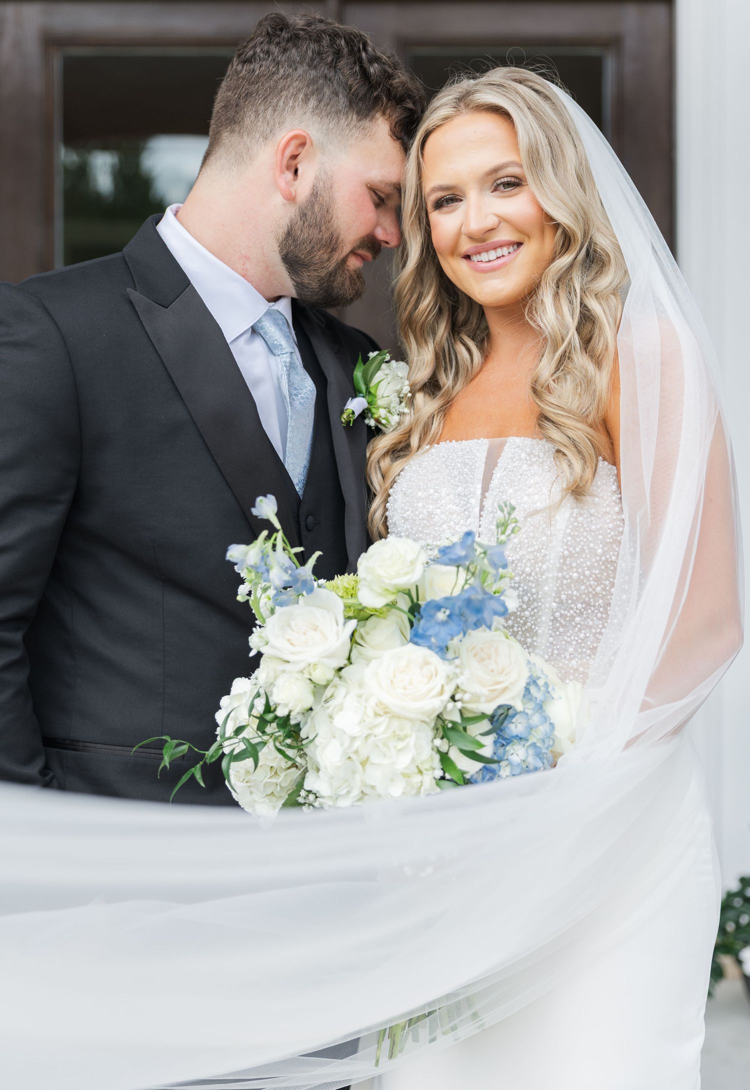Bride and groom portrait with white and blue hydrangea bouquet, veil flowing, groom in black suit.