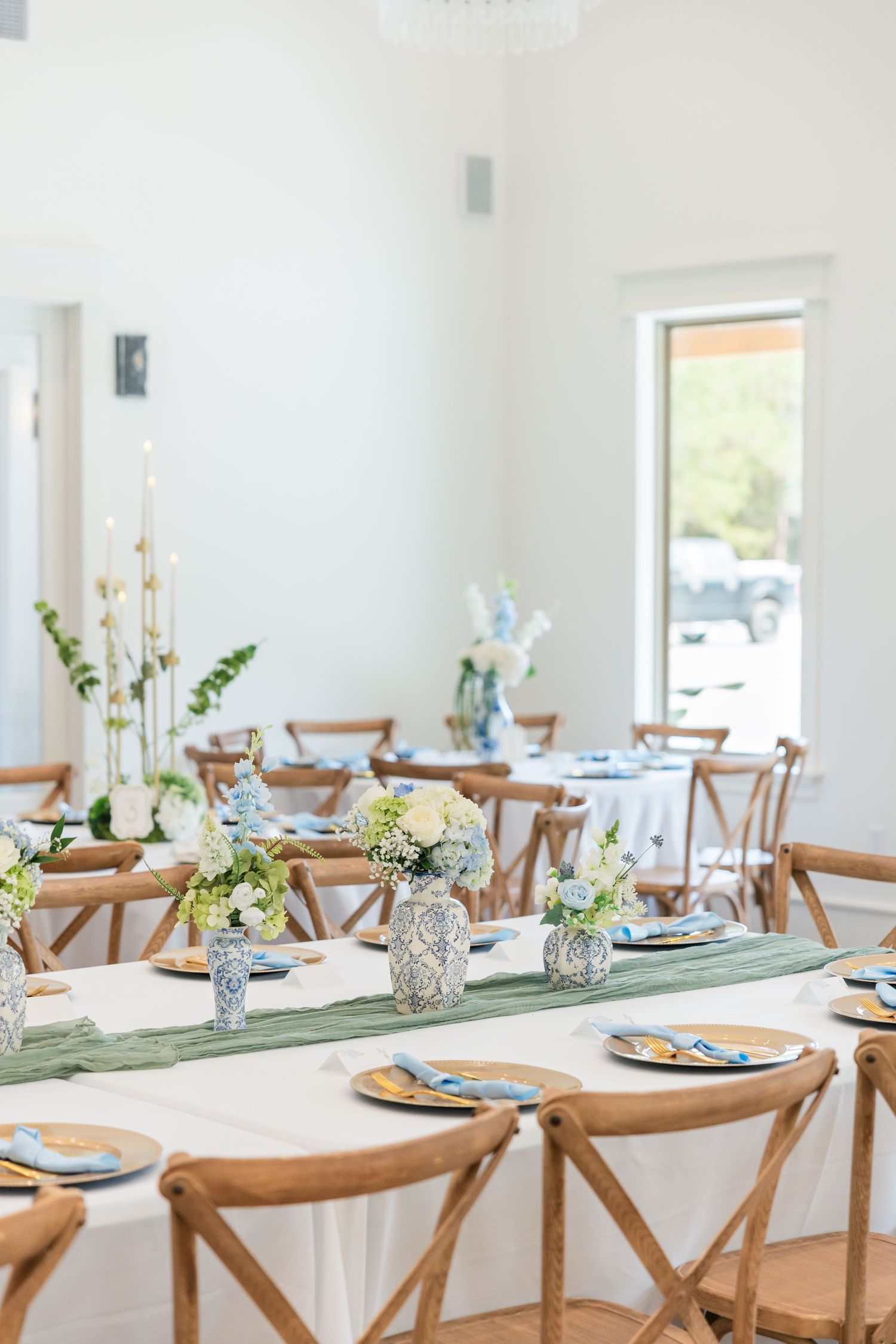 Elegant wedding reception tables with green runners, blue and white floral centerpieces, wooden cross-back chairs, and gold charger plates.
