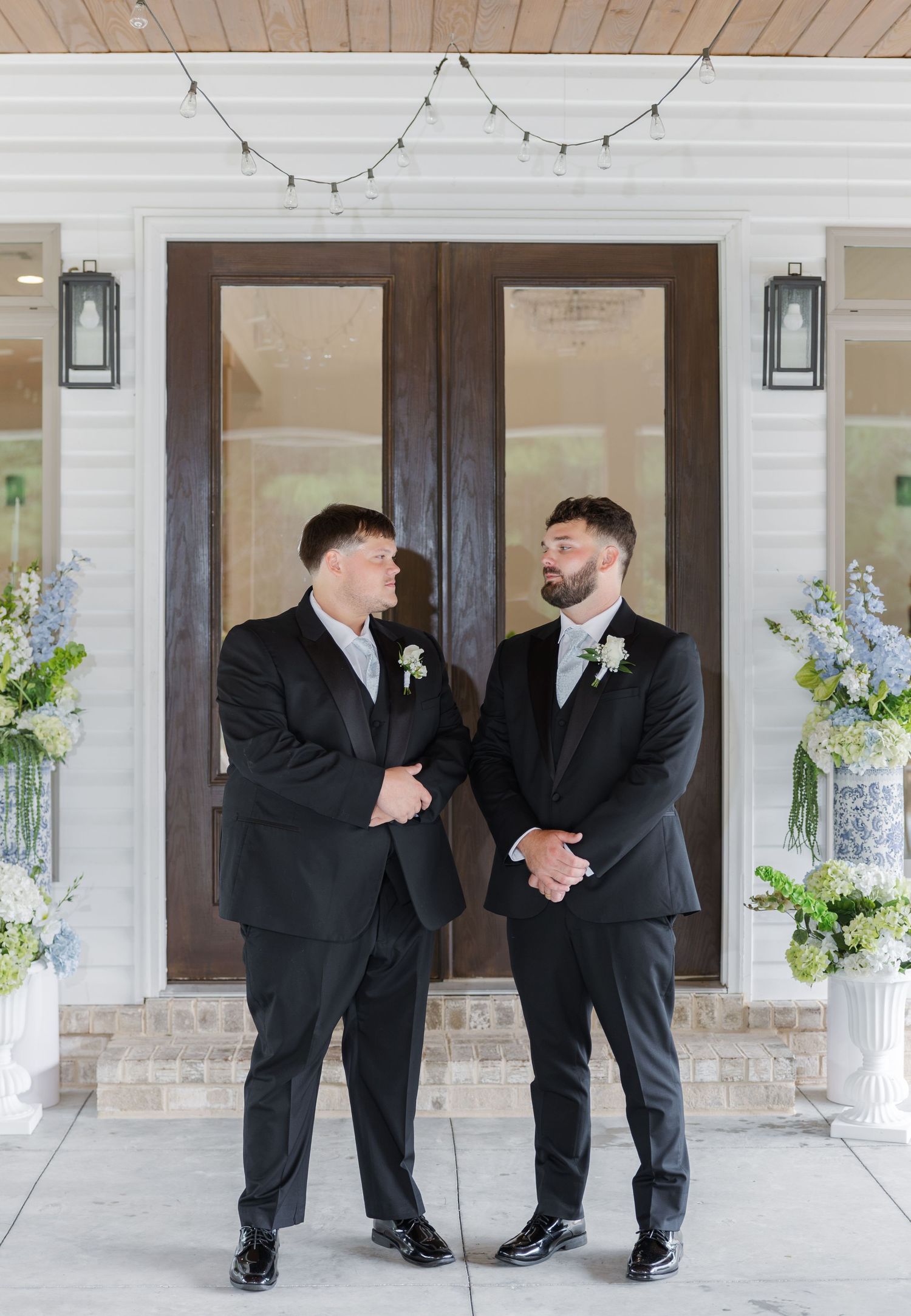 Two groomsmen in black suits with boutonnieres standing before elegant double doors flanked by floral arrangements.
