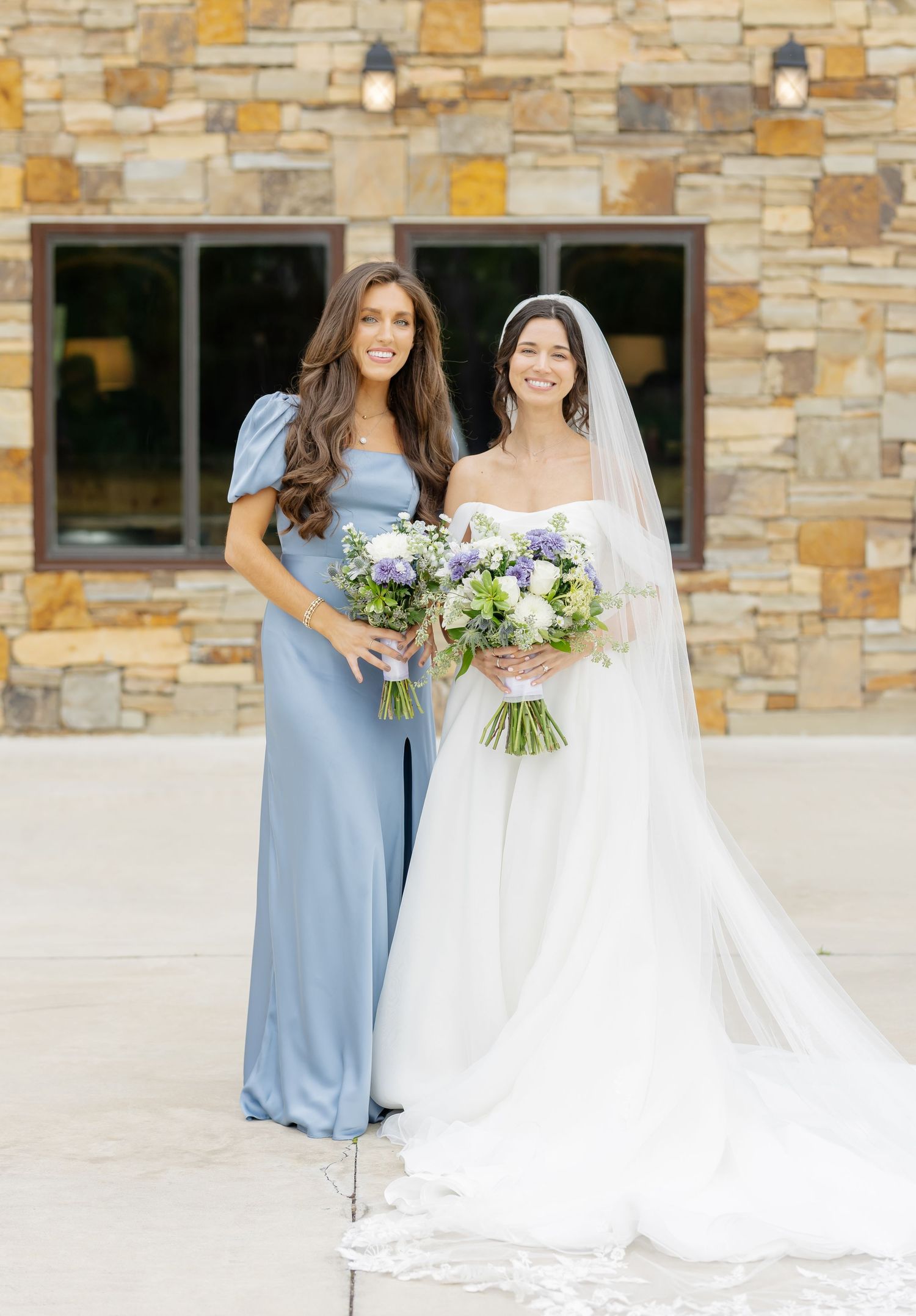 Bridesmaids in light blue dresses pose with bride in white gown against stone wall.