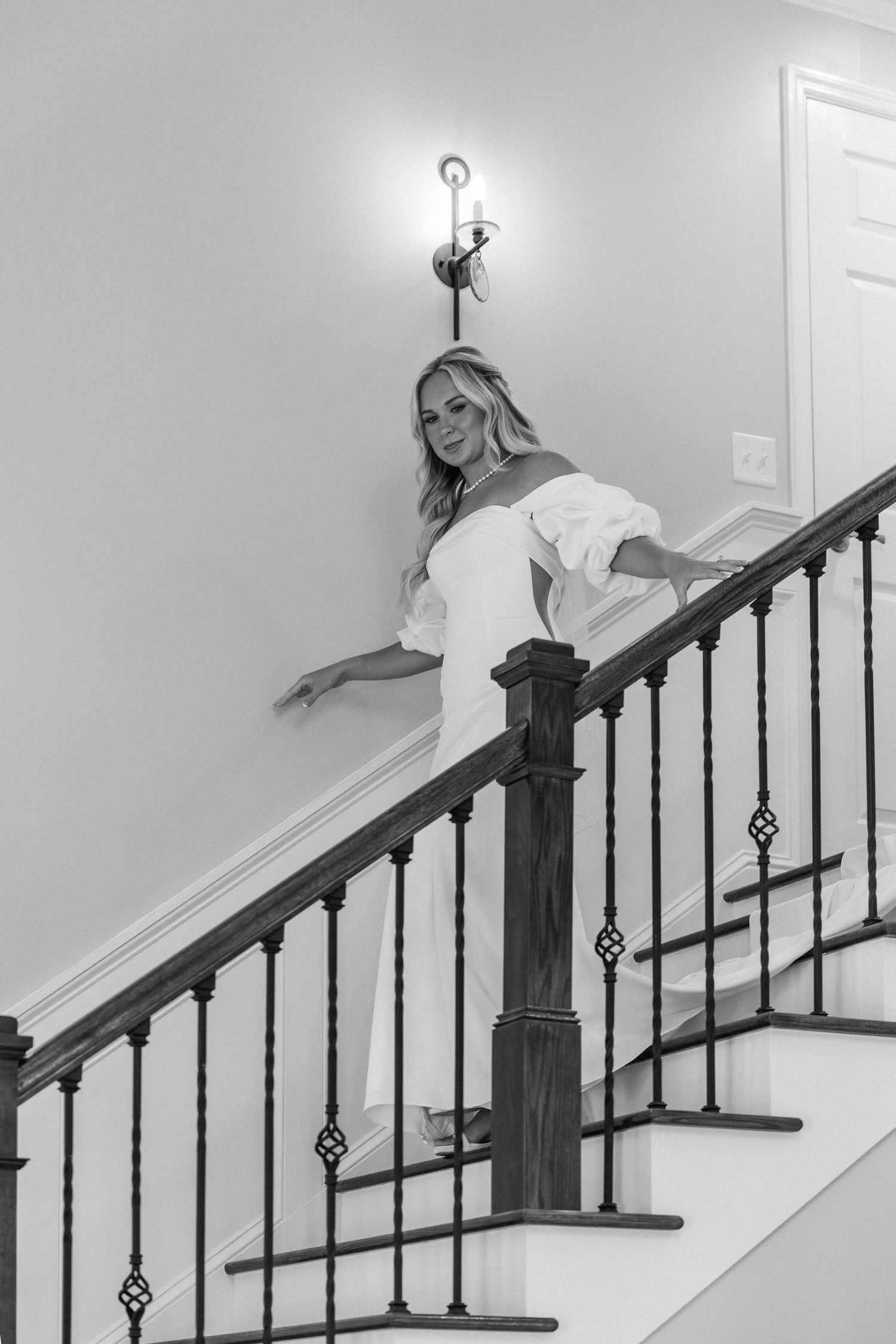 Black and white photo of blonde bride in off-shoulder gown posing gracefully on an elegant staircase.