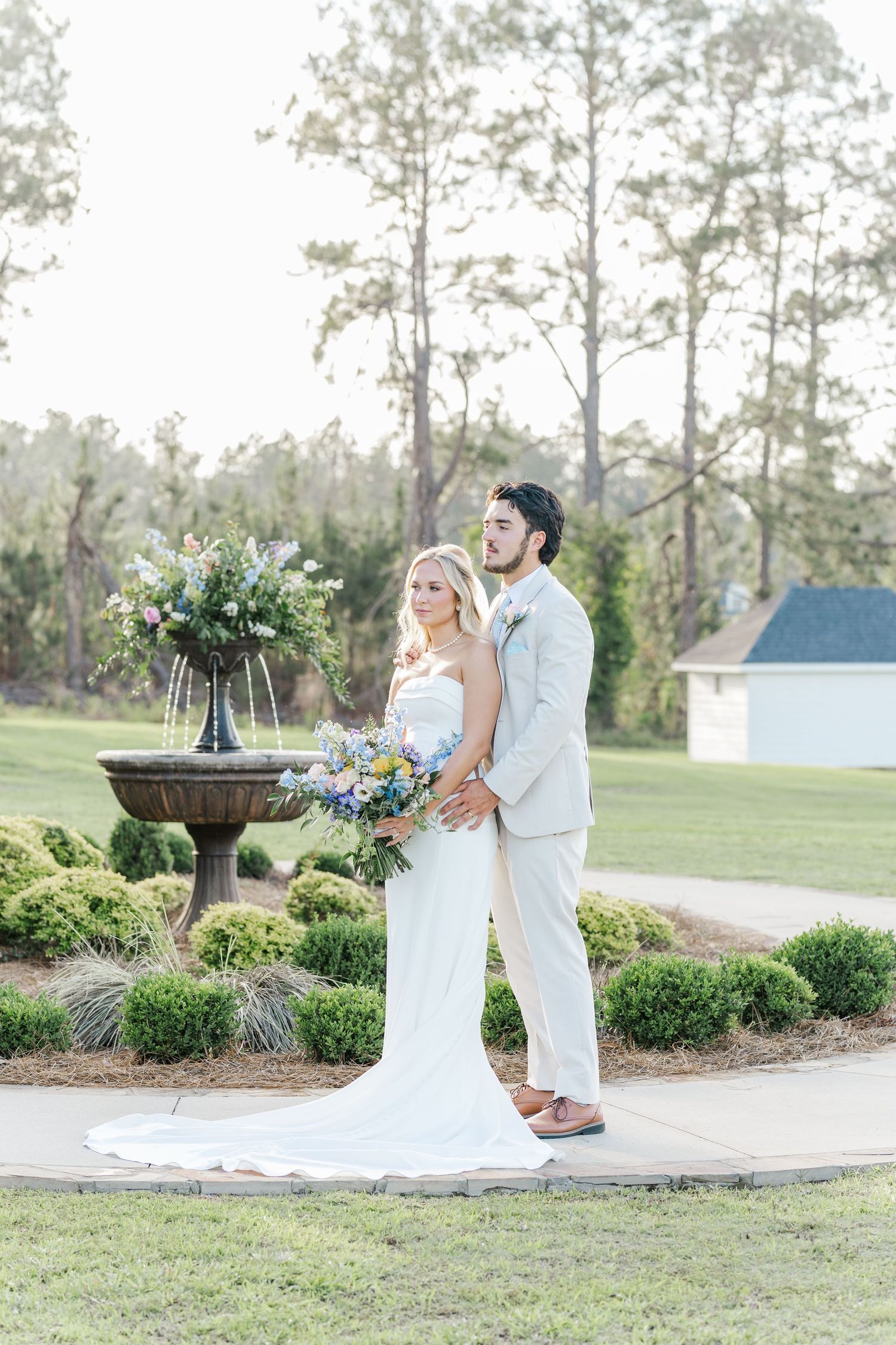 A bride and groom pose elegantly by a floral fountain in a lush garden, she holding a colorful bouquet.