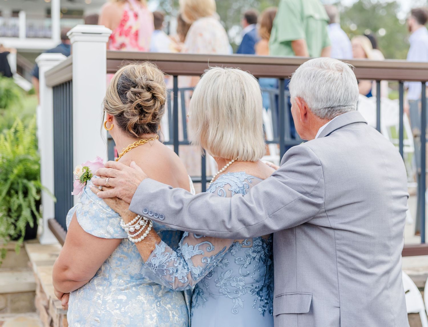 Couple in light blue lace and gray suit embracing from behind while watching outdoor wedding ceremony.
