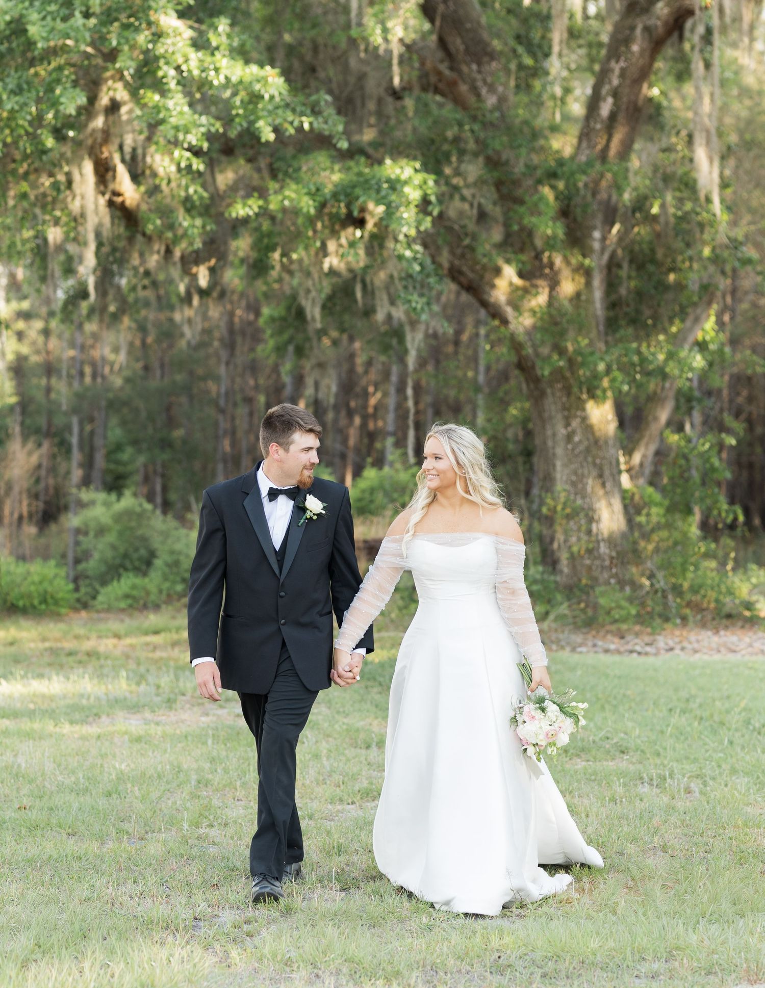 Bride and groom walking hand in hand through lush green grounds with Spanish moss-draped oak trees.