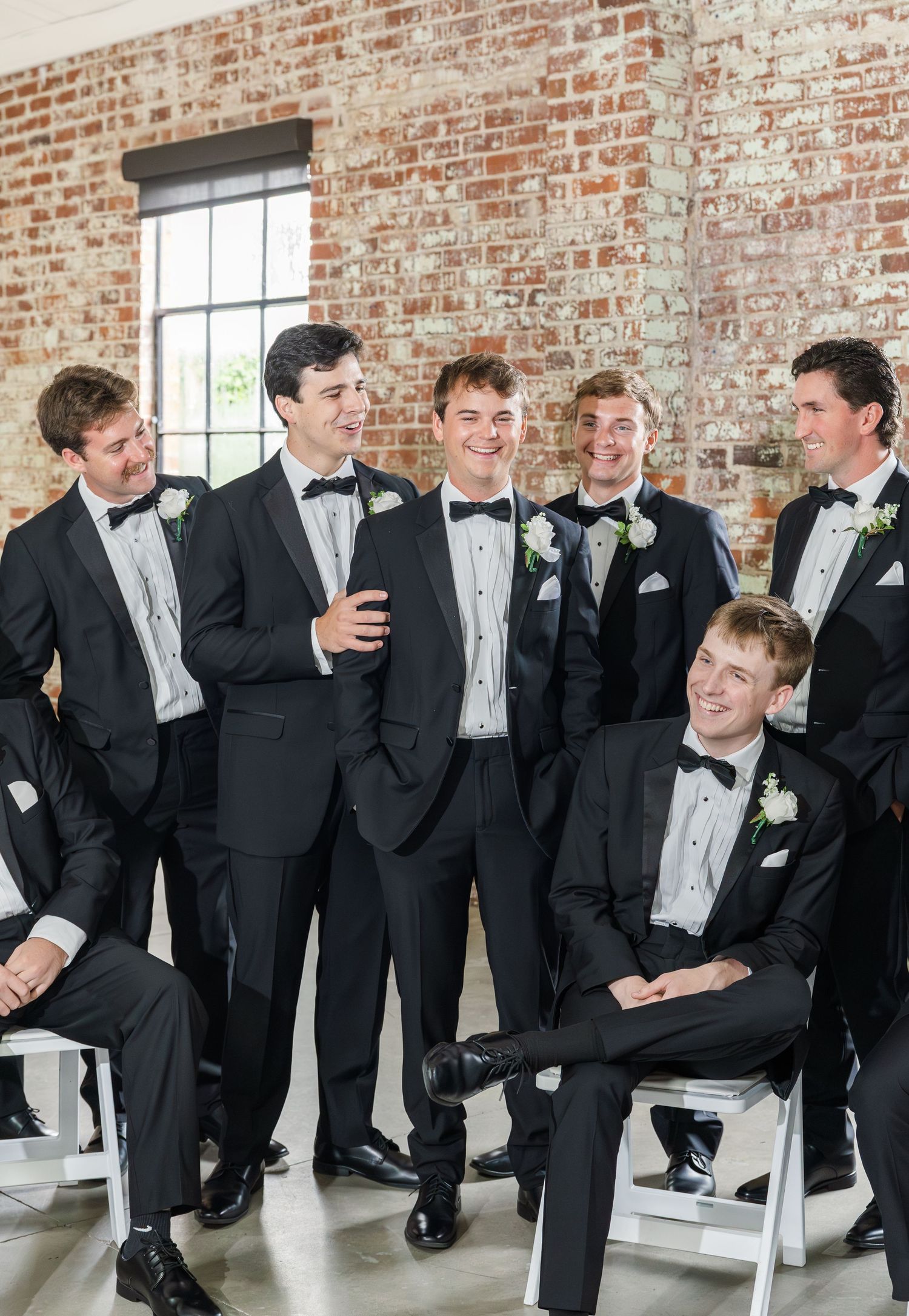 Six groomsmen in black tuxedos with bow ties laughing together against a rustic exposed brick wall.