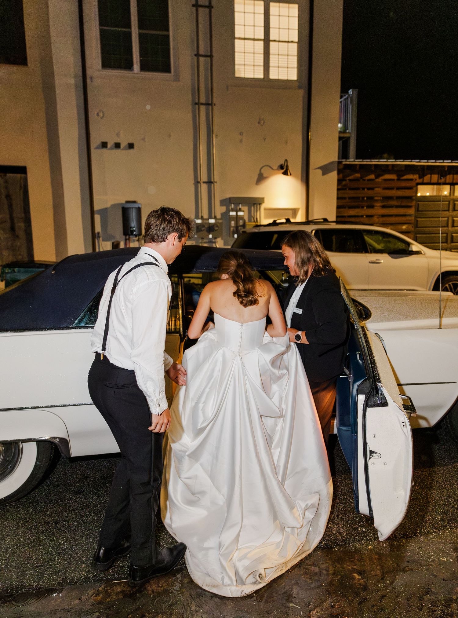 Bride in silver gown entering white vintage car with groom and helper after wedding reception.