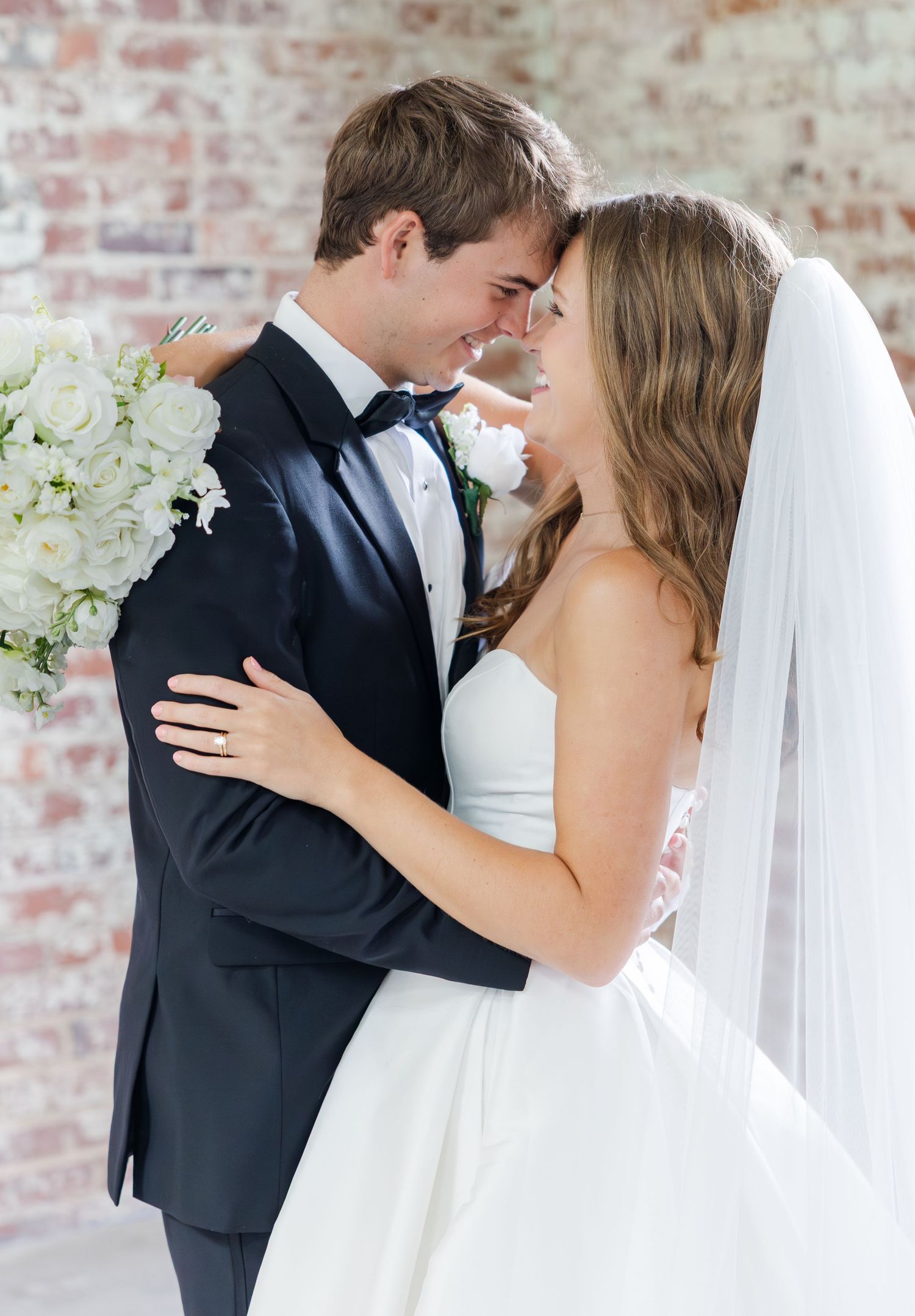 Bride and groom share an intimate forehead touch, she holds white bouquet wearing strapless gown with veil.