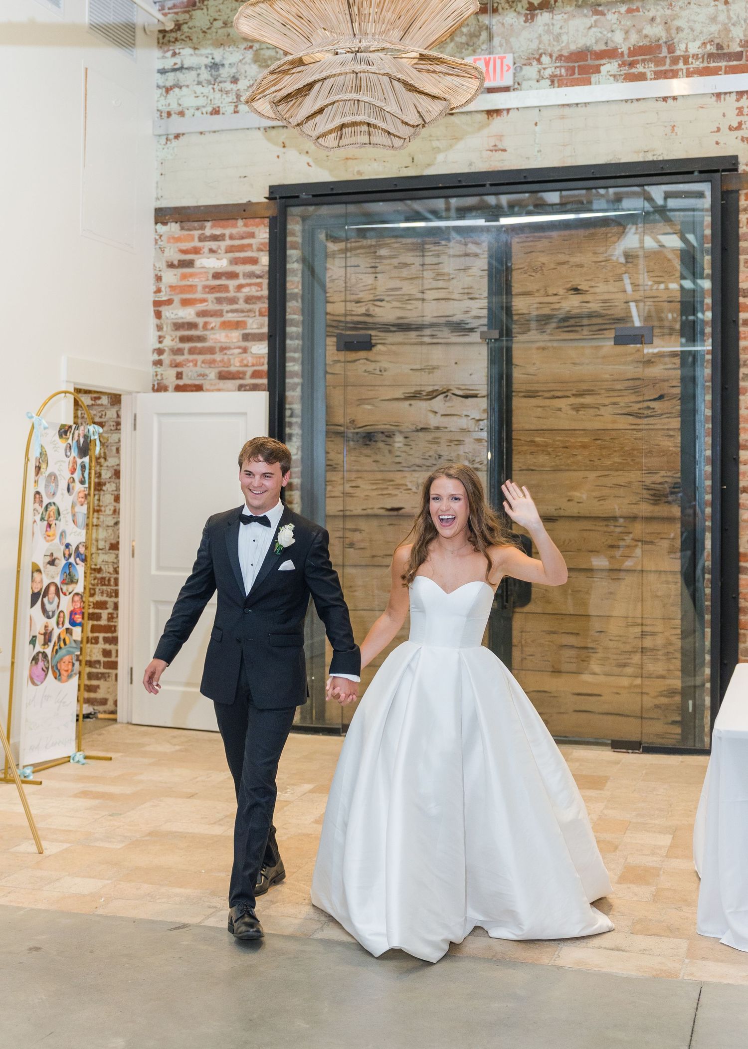 Smiling bride and groom enter wedding reception holding hands at industrial brick venue.