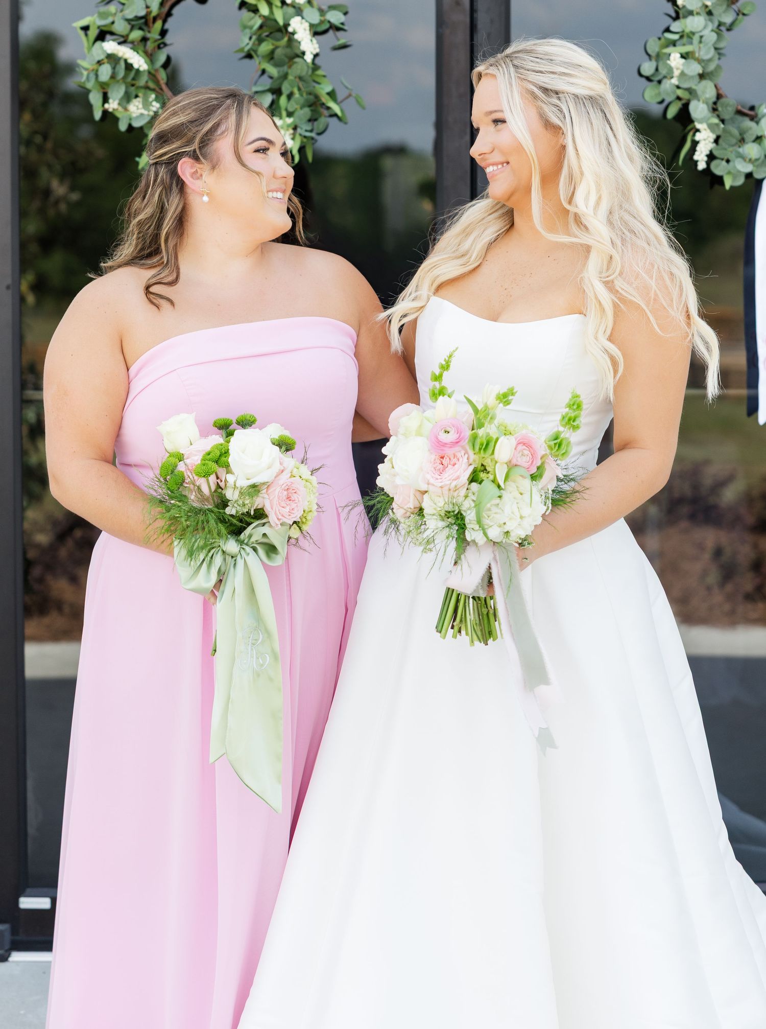 Bride in white strapless gown and bridesmaid in pink dress holding floral bouquets at wedding.