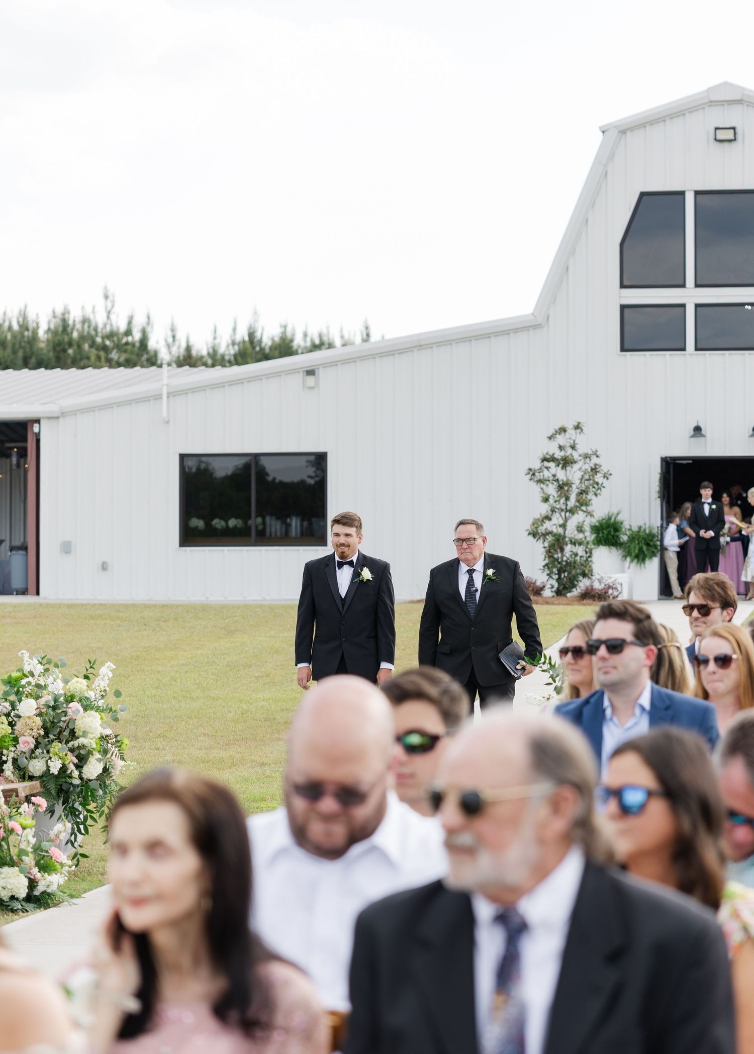 Groom walks down outdoor wedding aisle toward seated guests with white barn venue in background.