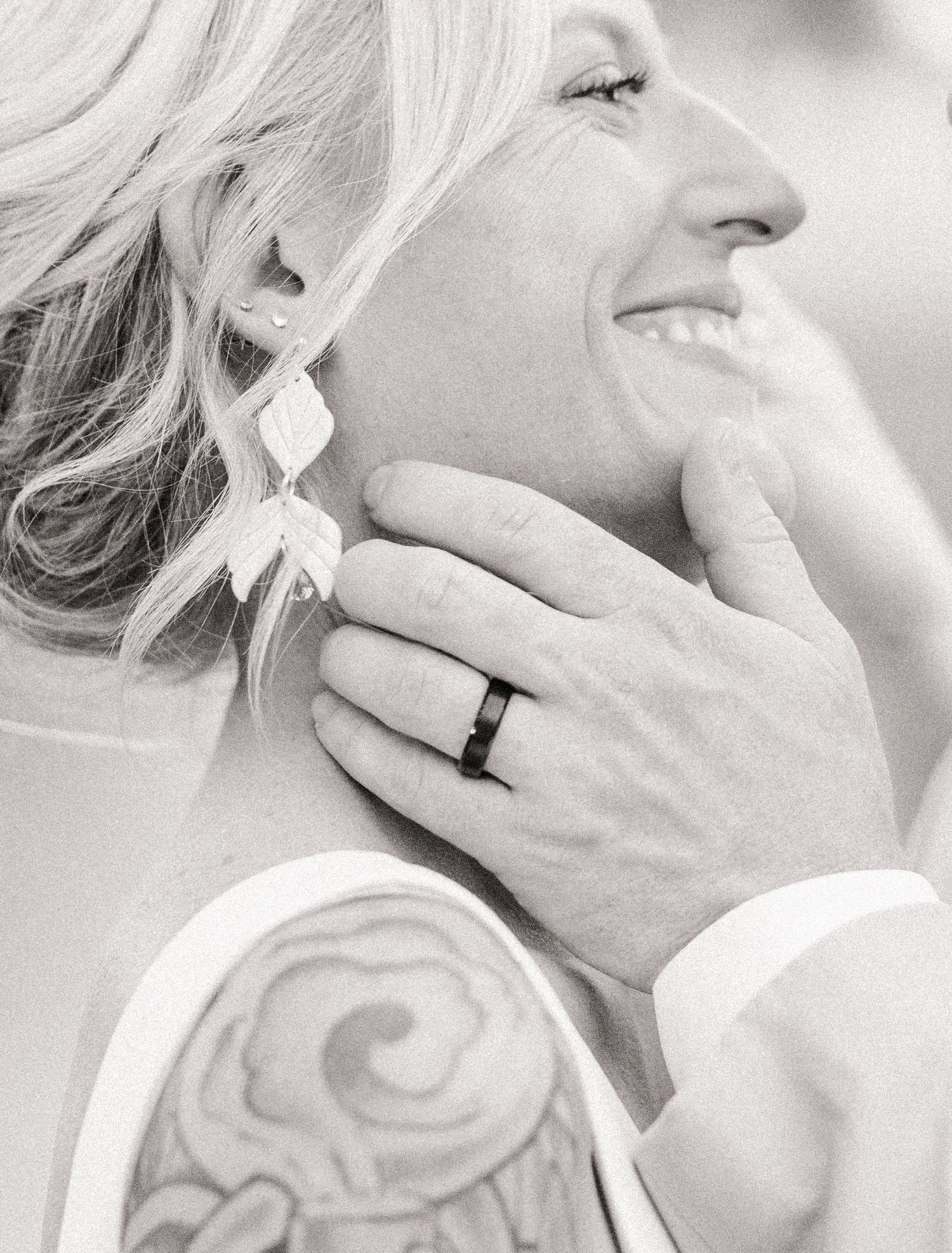 Black and white close-up of smiling bride with groom's tattooed hand gently touching her face.