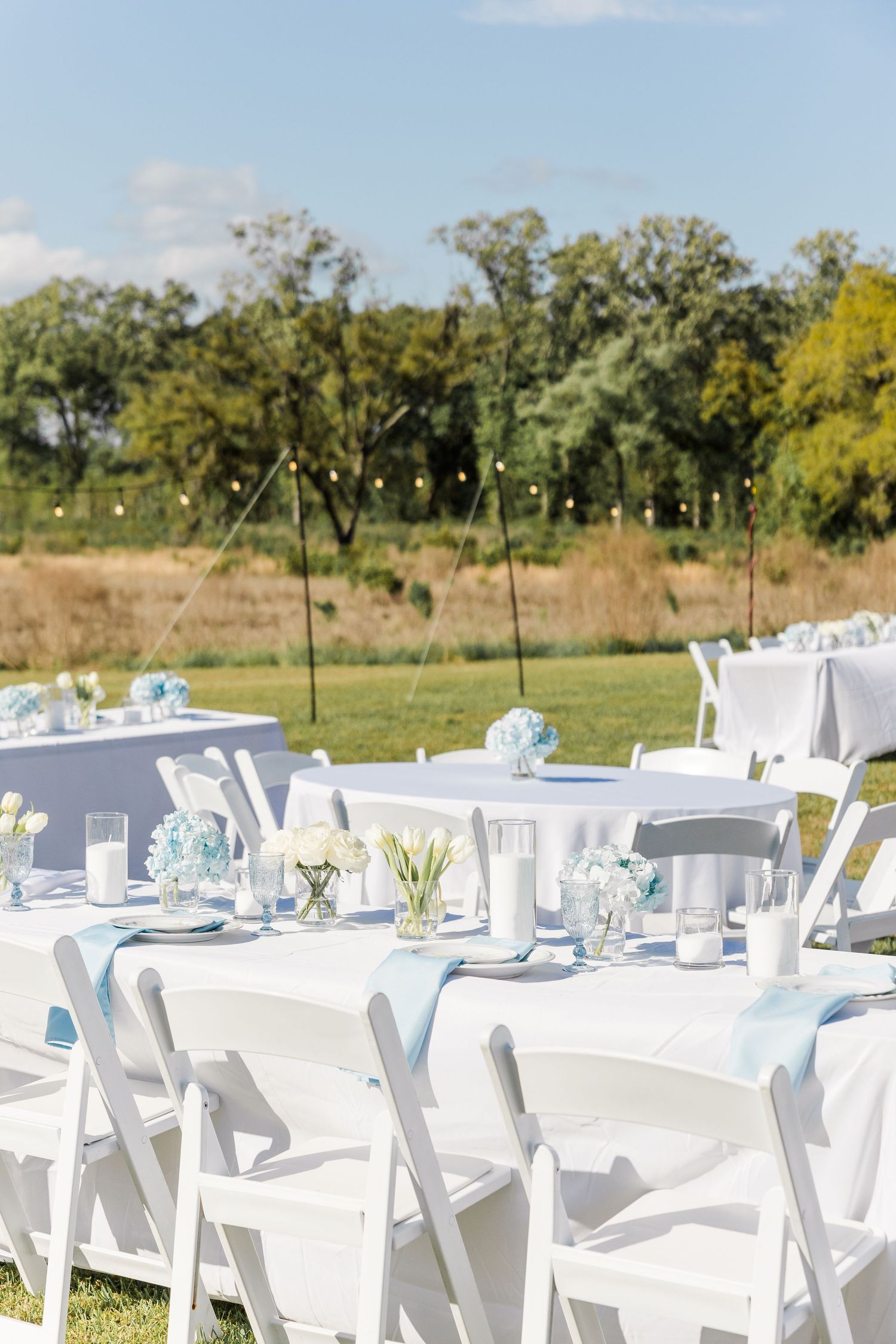 Outdoor wedding reception tables set with white linens, light blue runners, floral centerpieces, and white chairs under blue sky.