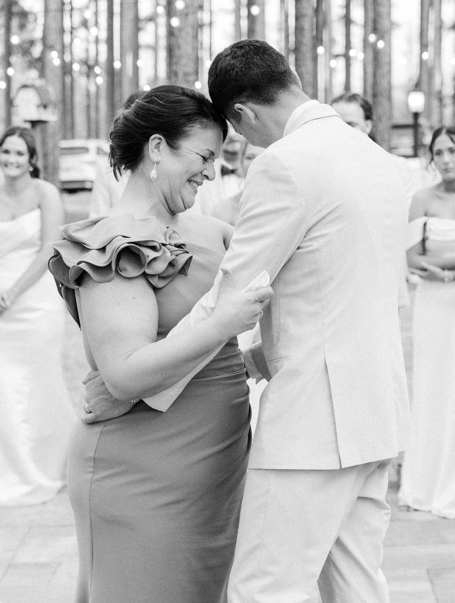 Black and white photo of groom in white suit sharing mother-son dance at wedding reception.