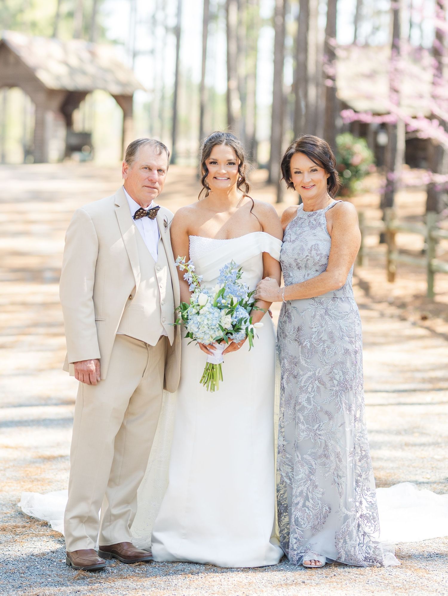Bride in a white strapless gown poses with her parents, holding a blue hydrangea bouquet, surrounded by pine trees.