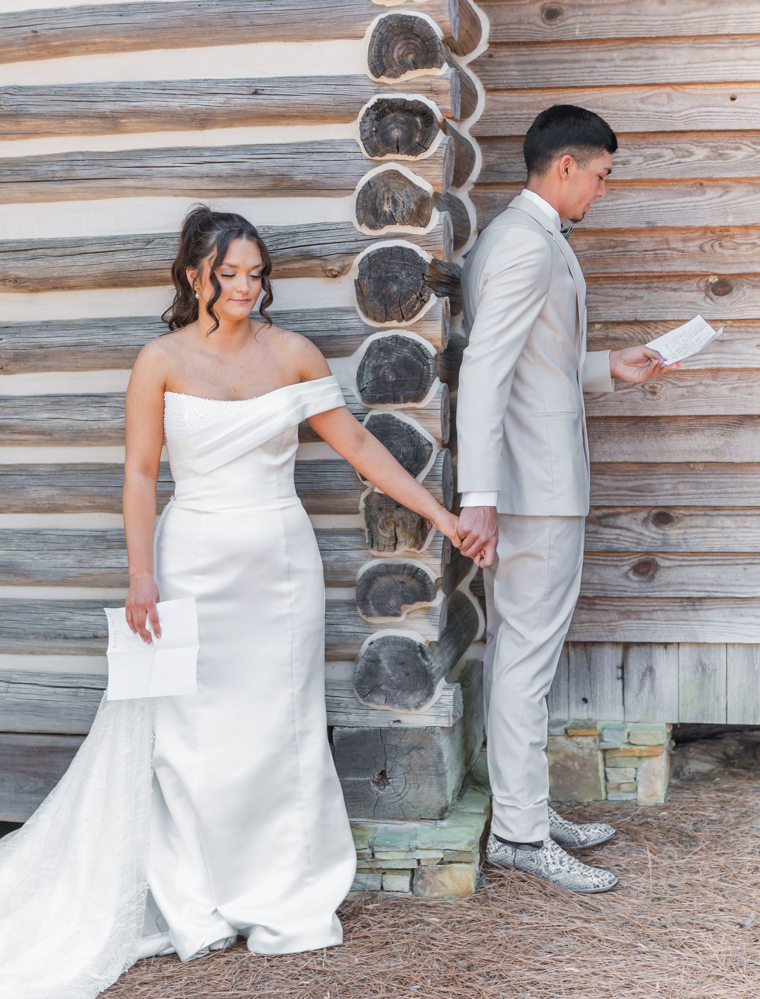 Bride and groom share a private vow reading moment, holding hands beside a rustic log cabin wall.