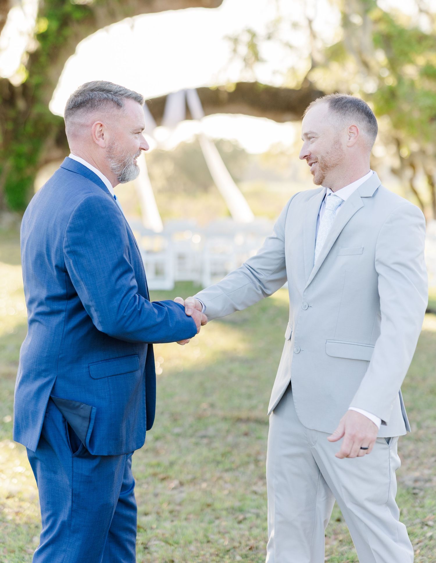 Two men in suits shake hands warmly at an outdoor wedding venue with white chairs in background.