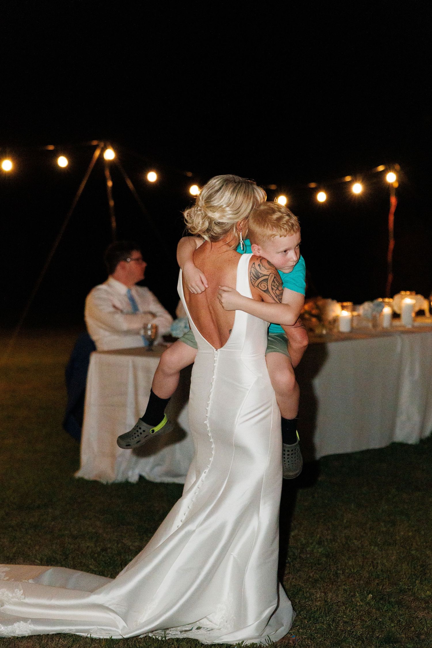 Bride in backless white gown holds young child during outdoor evening wedding reception under string lights.