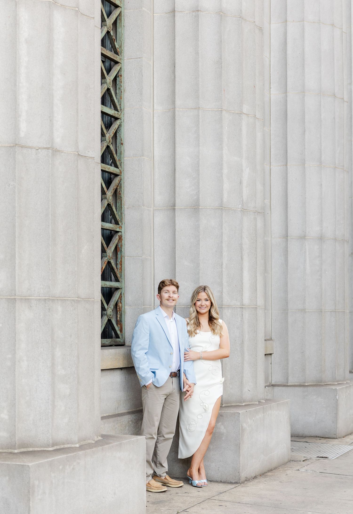 Couple posing by large stone columns, man in light blue blazer and woman in white dress, elegant outdoor portrait.