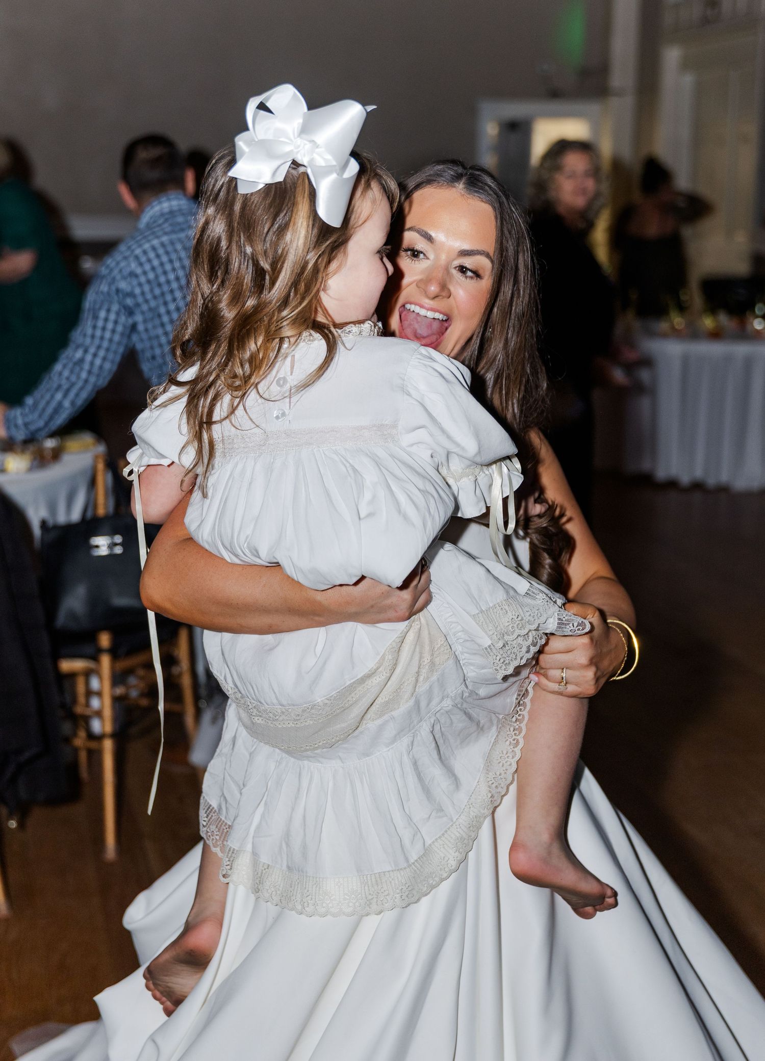 Bride in white gown joyfully hugging barefoot flower girl on wedding reception dance floor.