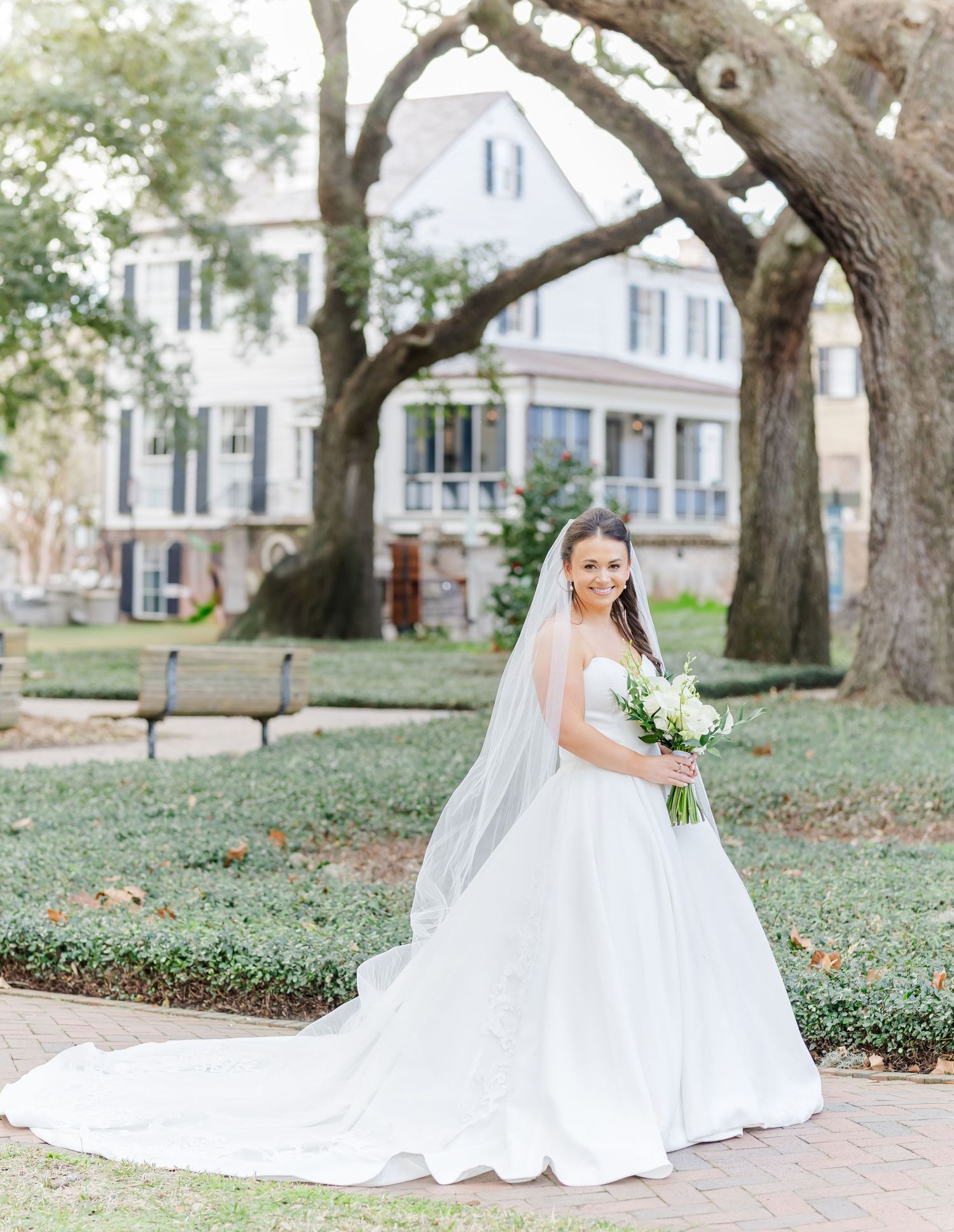 Bride in a flowing white gown with veil holds bouquet in a lush garden with a historic white mansion backdrop.