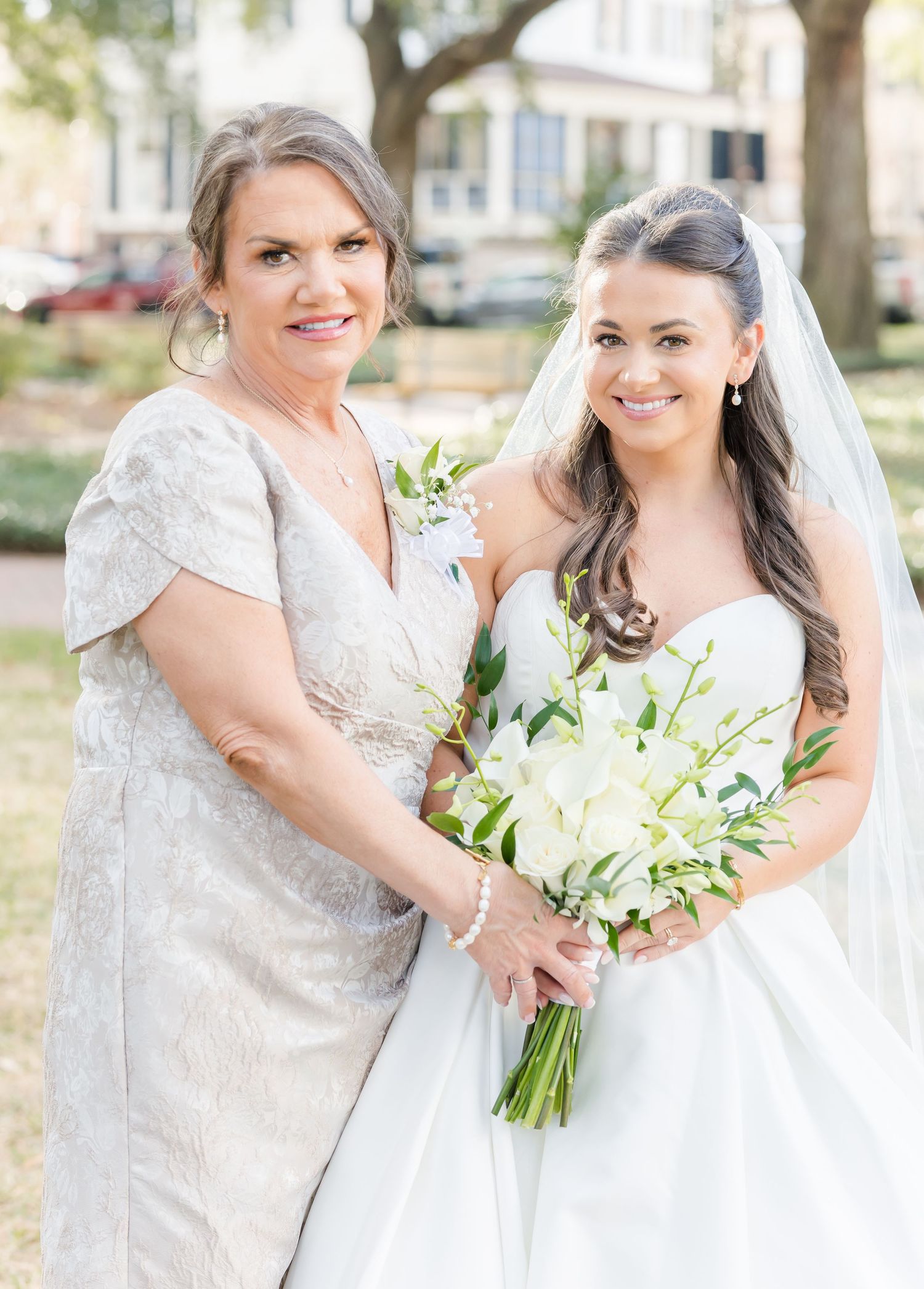 Bride in strapless white gown holding white floral bouquet poses with mother in silver dress.