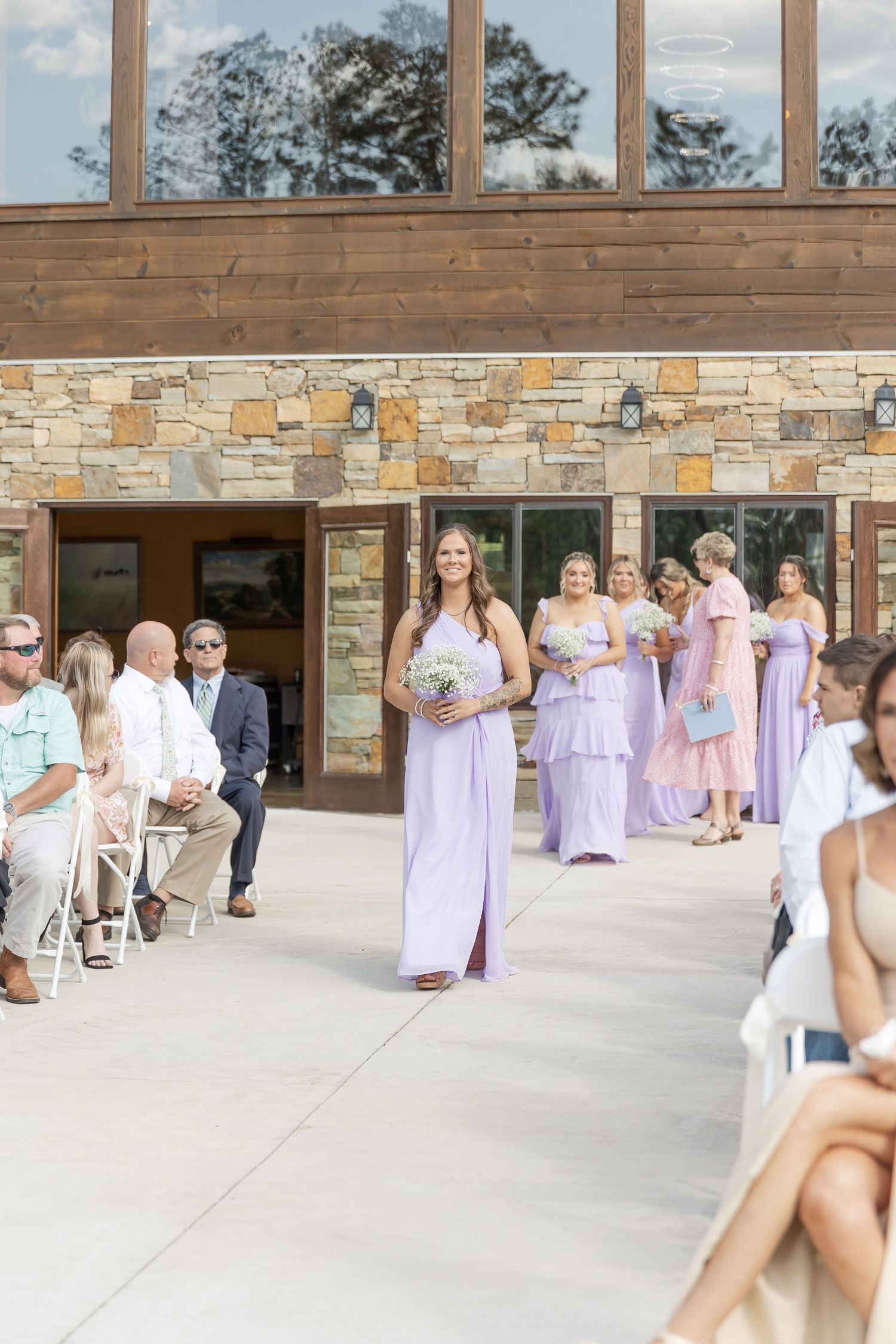 Bridesmaid in flowing lavender dress walks down aisle at stone and wood venue.