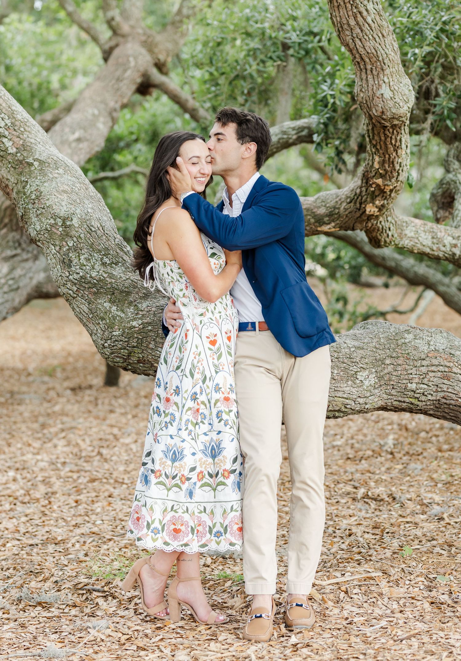 Couple embracing by a large oak tree, man kissing woman's cheek, she wears a floral dress, he wears a navy blazer.