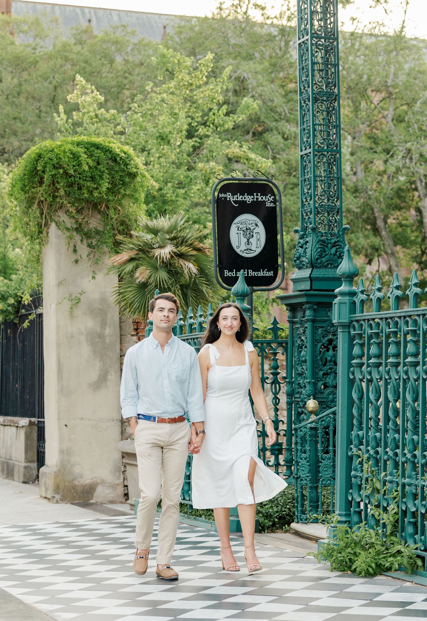 Couple walking hand in hand past teal iron gates and a bed and breakfast sign on a charming cobblestone street.