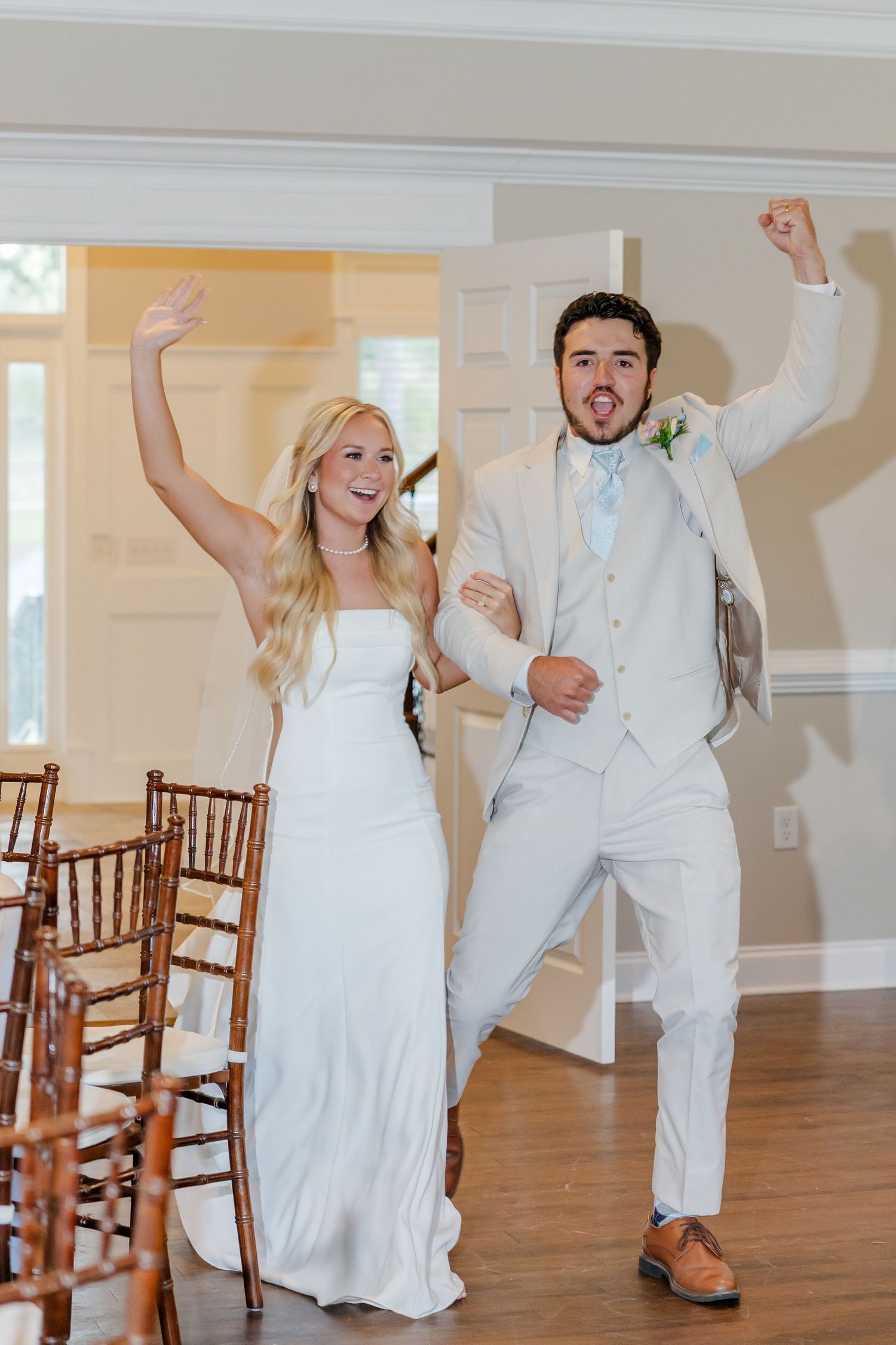 A joyful bride in a white gown and groom in a light suit celebrate their grand entrance with raised arms.