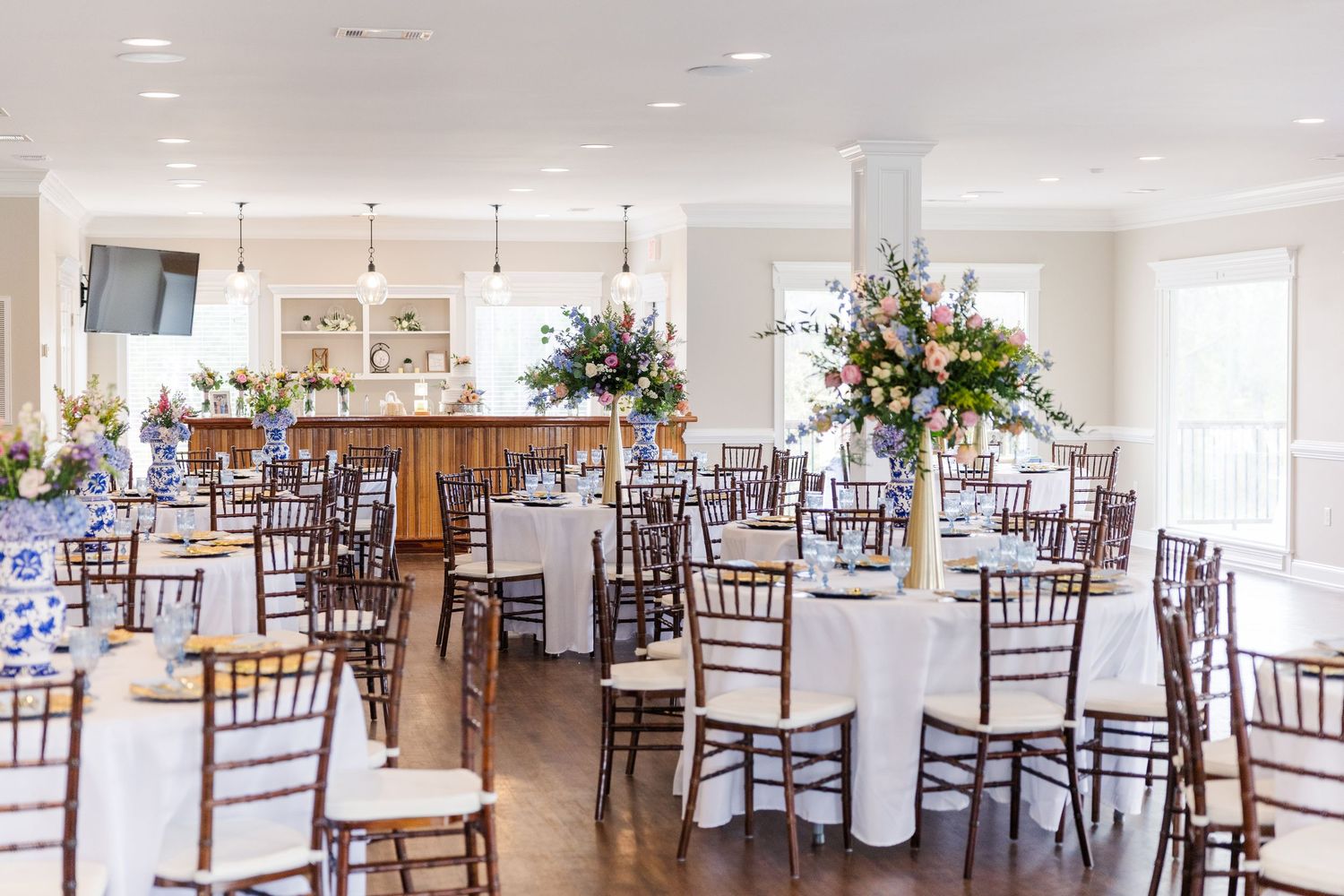 Elegant wedding reception hall with chiavari chairs, white linens, tall floral centerpieces, and bar.
