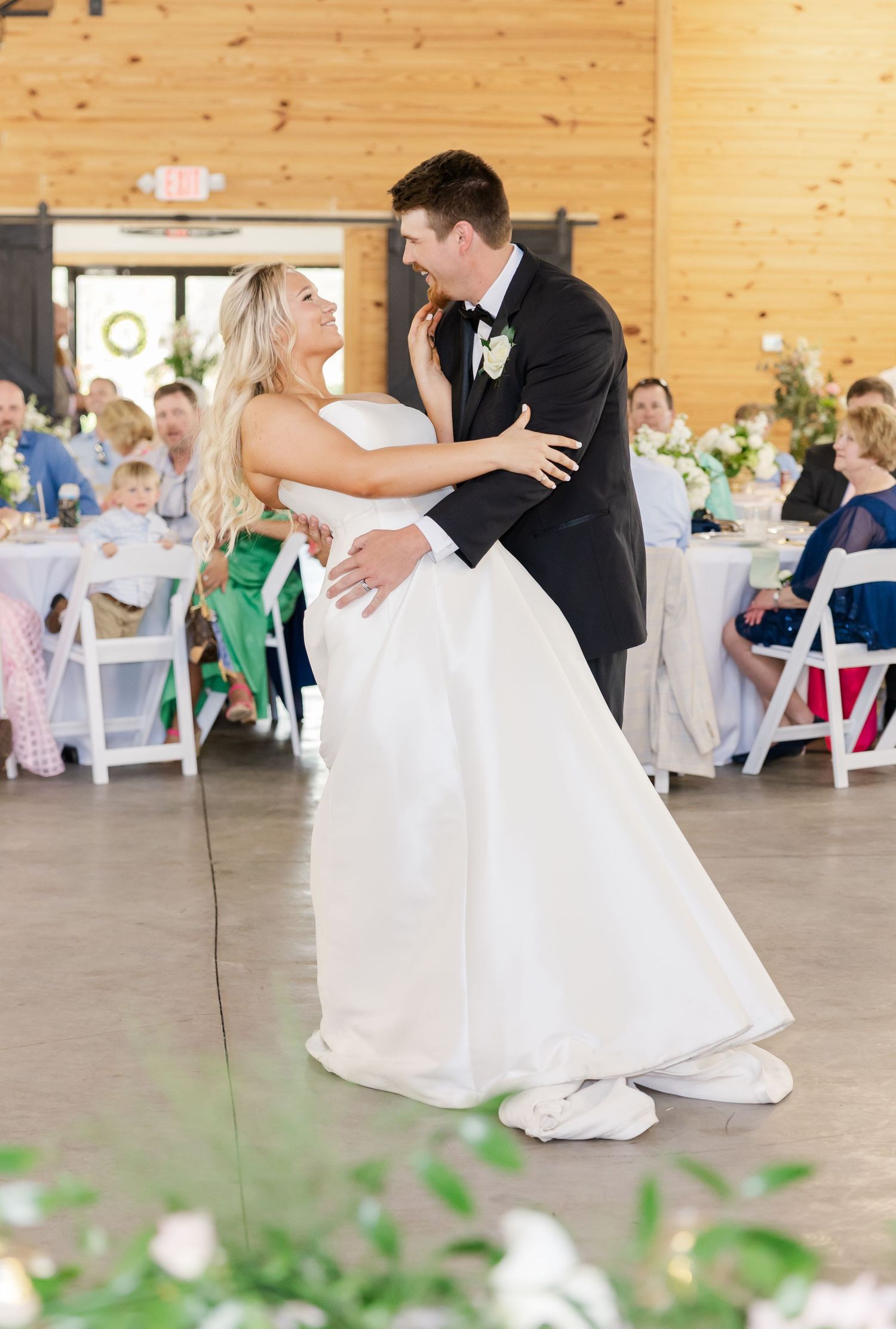 Bride and groom share romantic first dance in rustic barn reception venue with guests seated nearby.