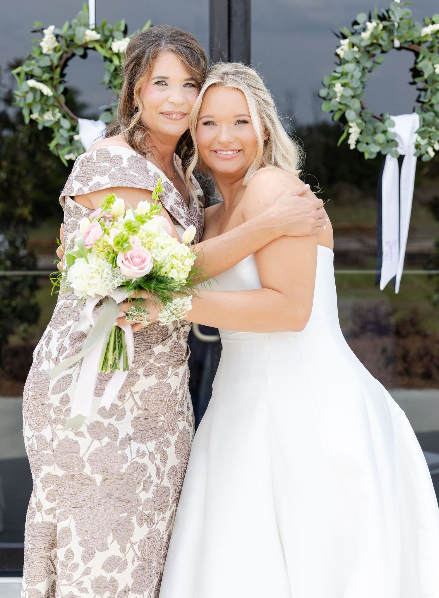 Bride in white strapless gown embraces woman in floral dress holding pink bouquet at outdoor wedding.