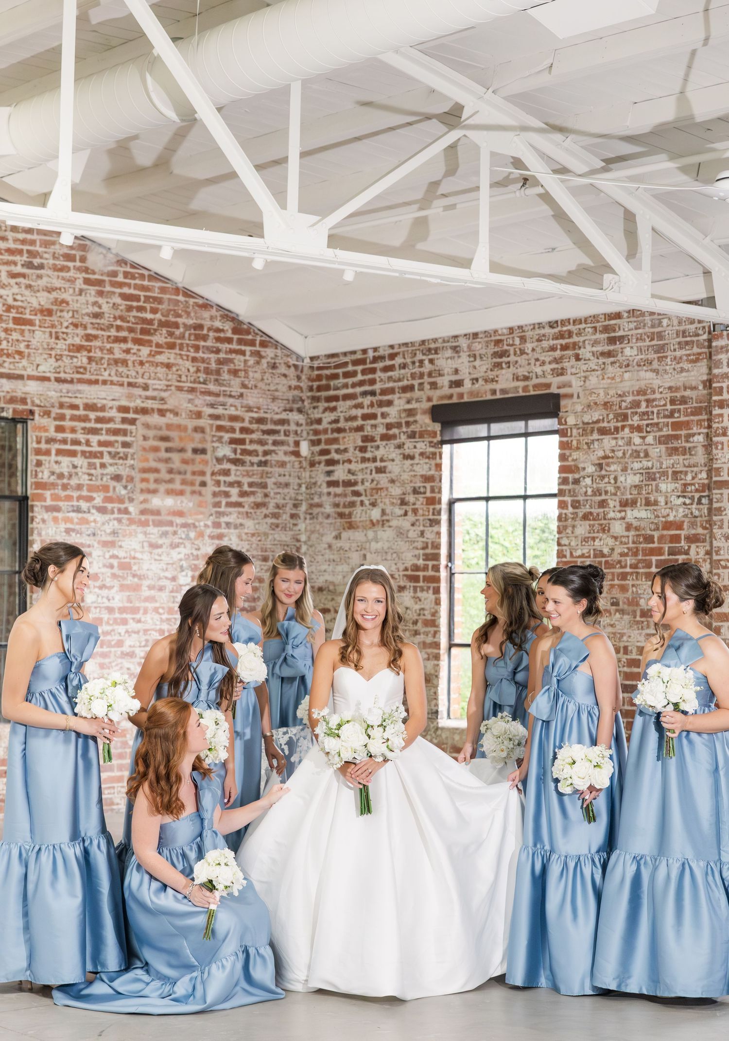 Bride in white ball gown surrounded by bridesmaids in blue dresses holding white bouquets indoors.