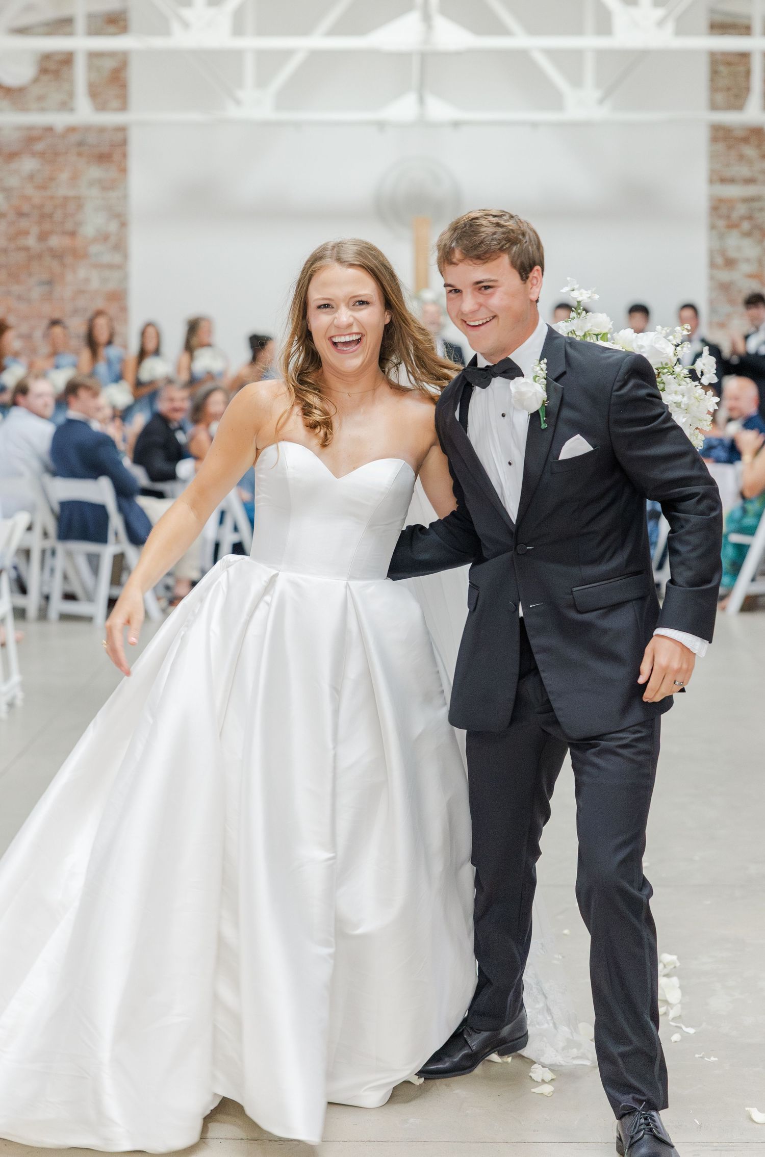 Laughing bride and groom exit wedding ceremony together in sunlit industrial venue with brick walls.