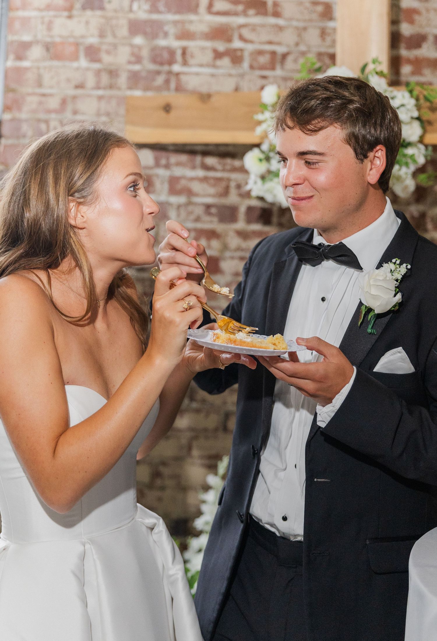 Bride and groom share wedding cake cutting moment, smiling together at rustic brick venue reception.