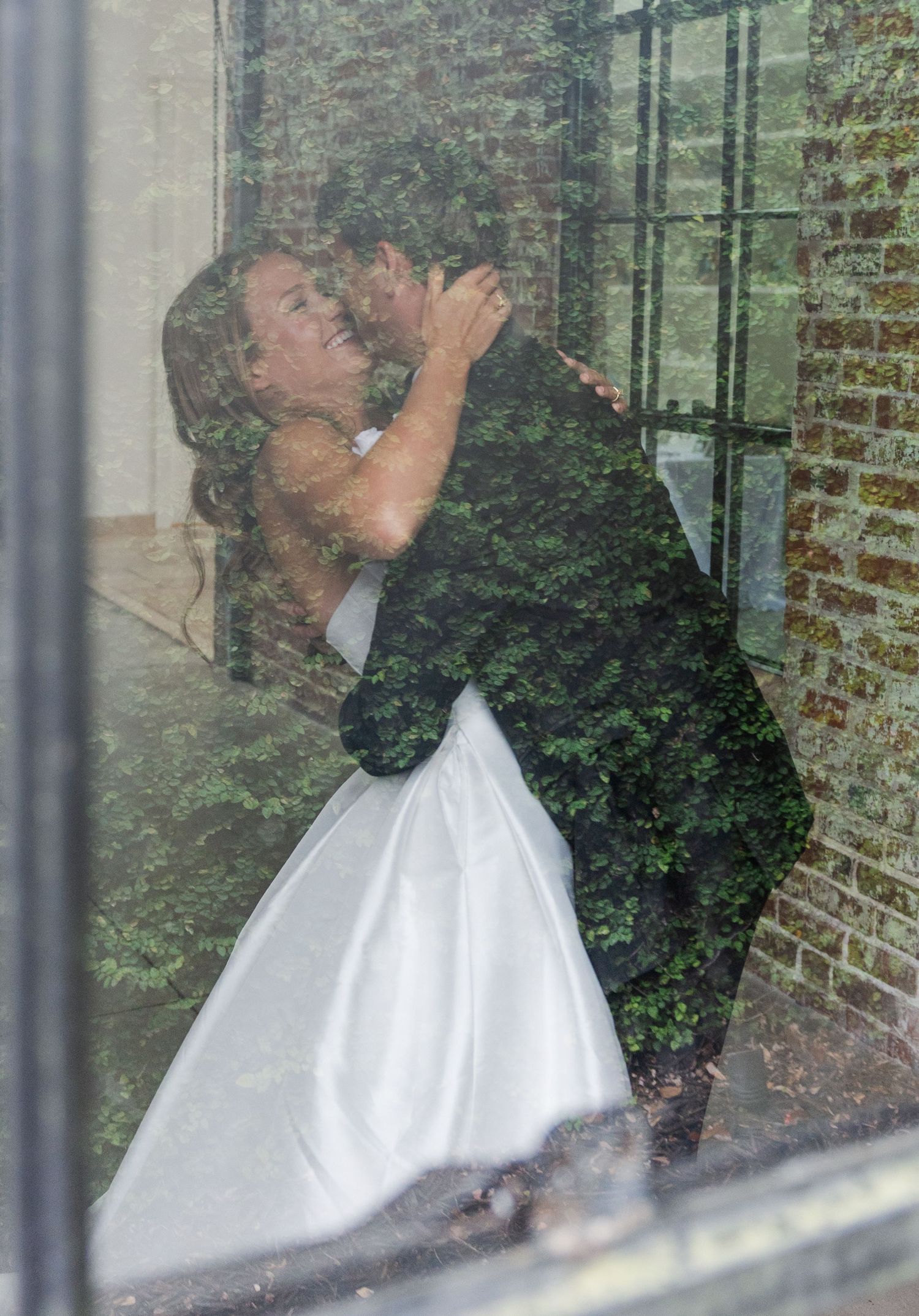 Newlyweds embrace in a passionate dip kiss photographed through a window with lush green garden reflections.