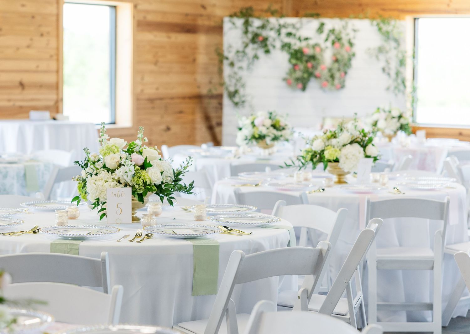 Elegant wedding reception hall with white round tables, green sage runners, white floral centerpieces, and wooden walls.