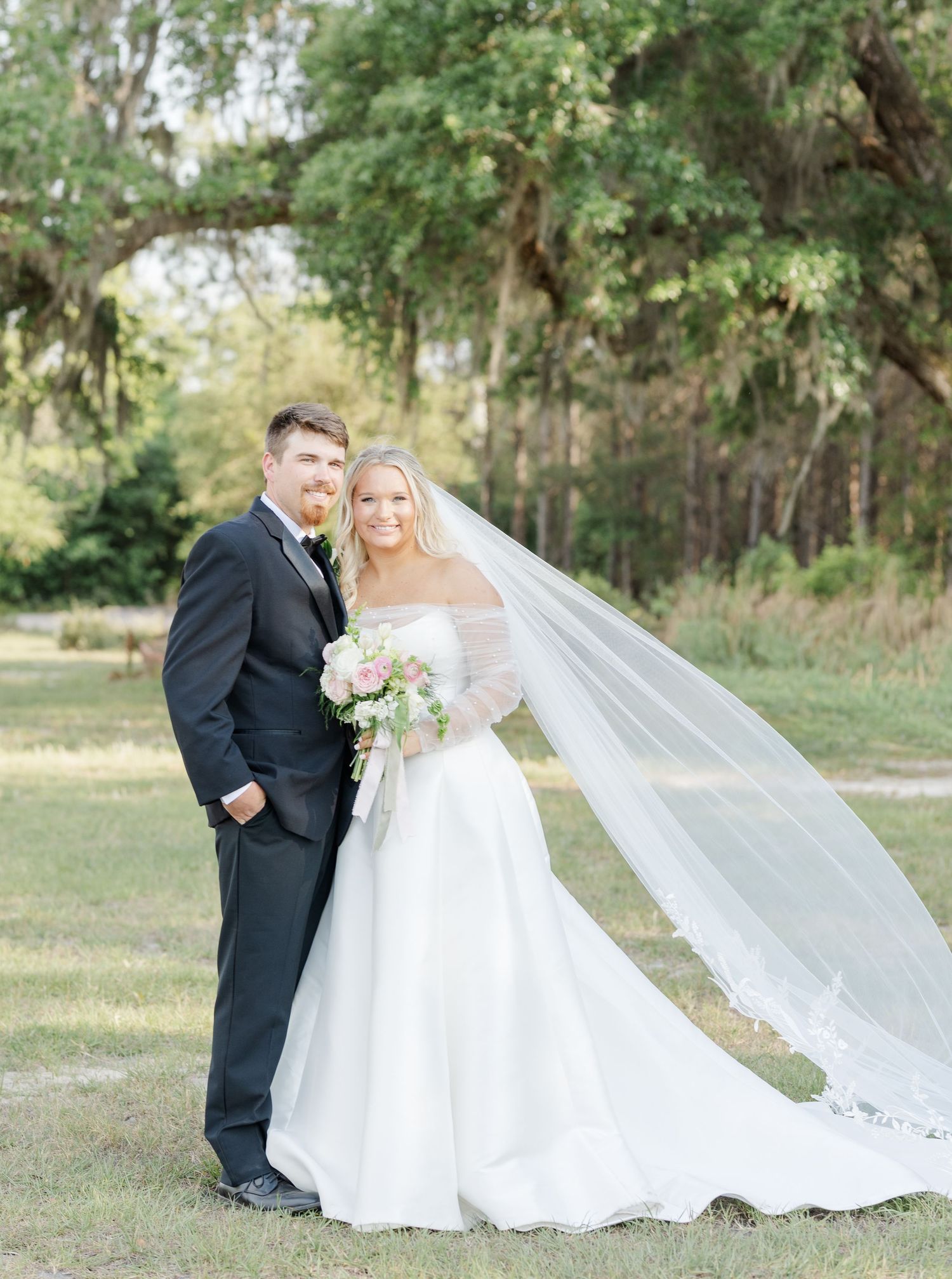 Bride and groom portrait outdoors with flowing cathedral veil and pink bridal bouquet under oak trees.
