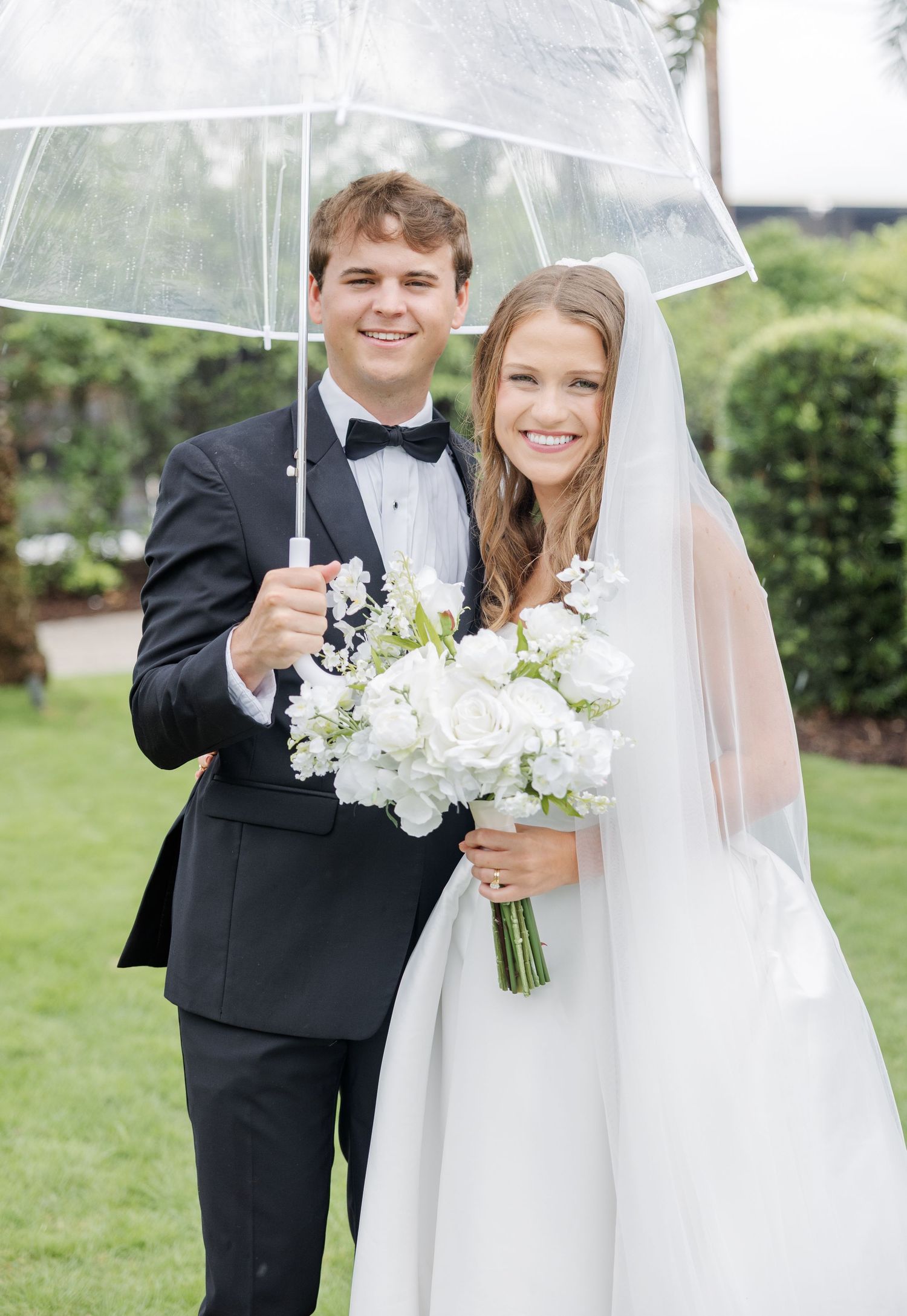 Smiling bride and groom posing under clear umbrella in rain, bride holding white rose bouquet in elegant wedding attire.