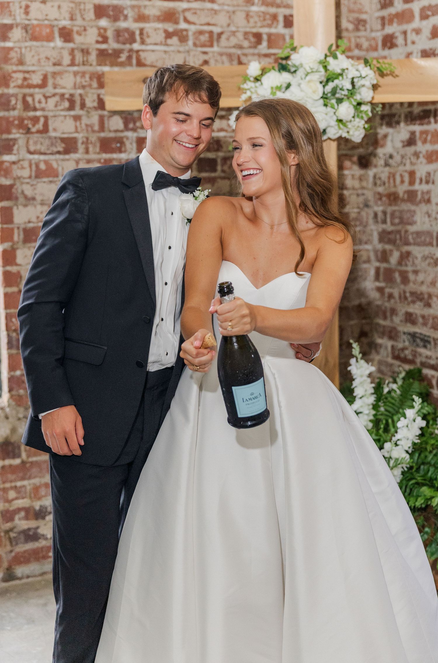 Newlyweds laugh joyfully while opening champagne bottle in front of floral arch at brick wall wedding venue.