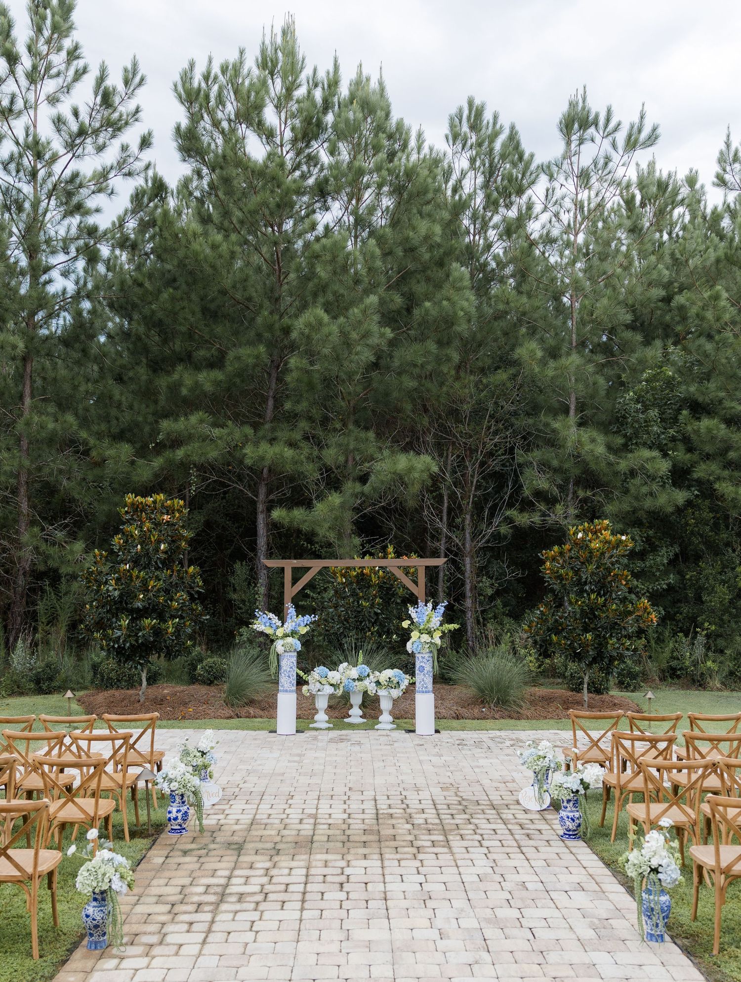 Outdoor wedding ceremony setup with wooden arch, blue and white floral arrangements, brick aisle, and wooden chairs amid pine trees.