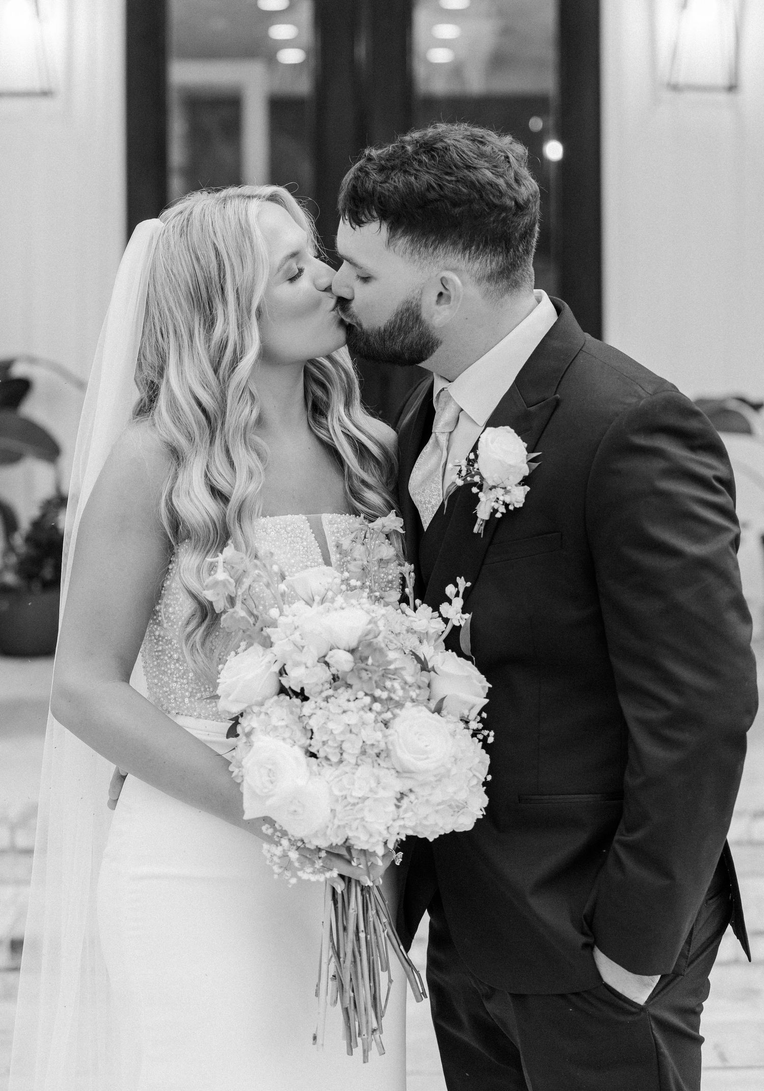 Black and white photo of a bride and groom sharing a kiss, bride holding a white floral bouquet.