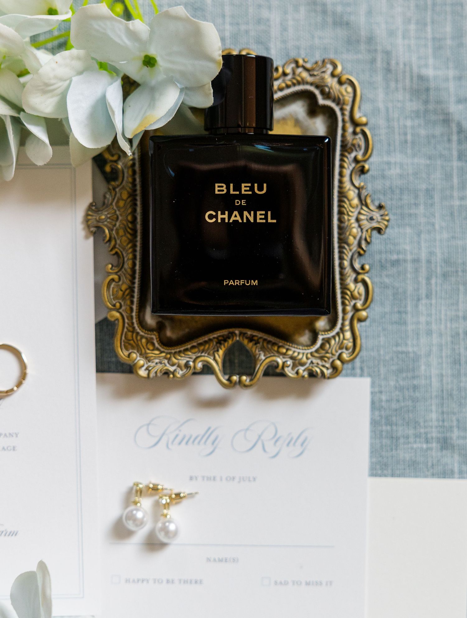 Bleu de Chanel parfum bottle on gold ornate tray beside pearl earrings and wedding invitation stationery.